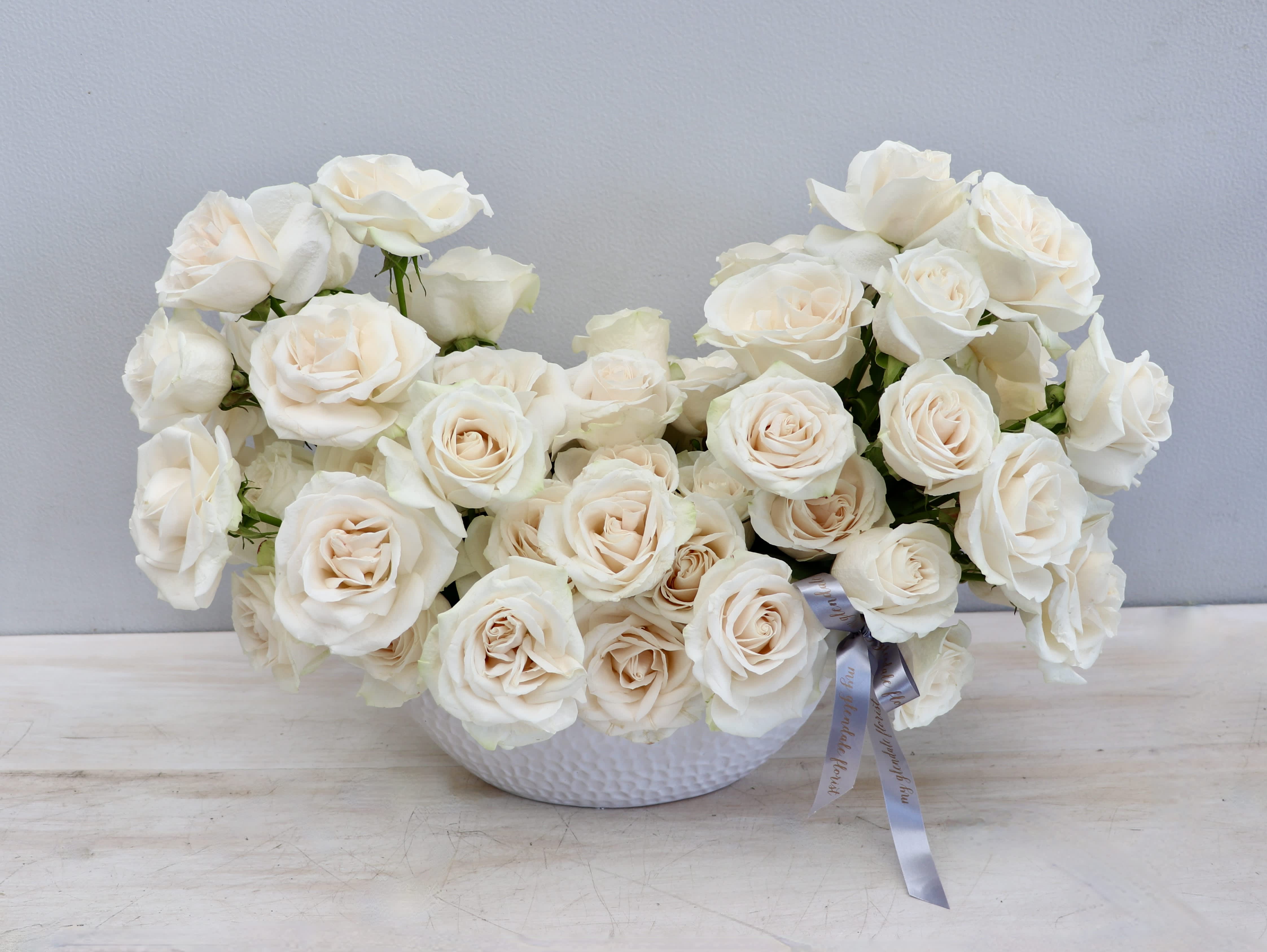 Low white bowl filled with cream roses and a gray ribbon accent