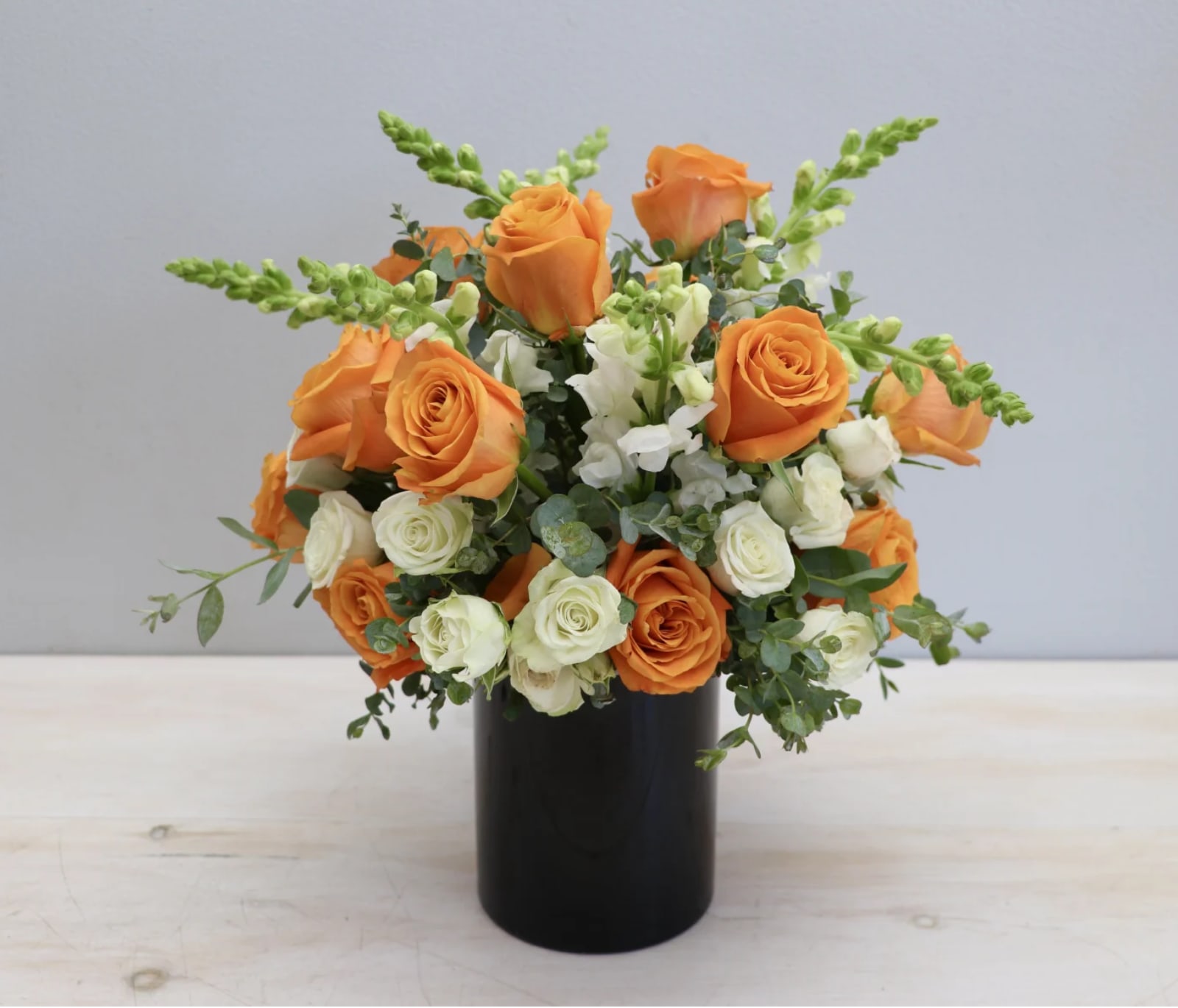 Orange and ivory roses with white snapdragons arranged in a black cylinder vase