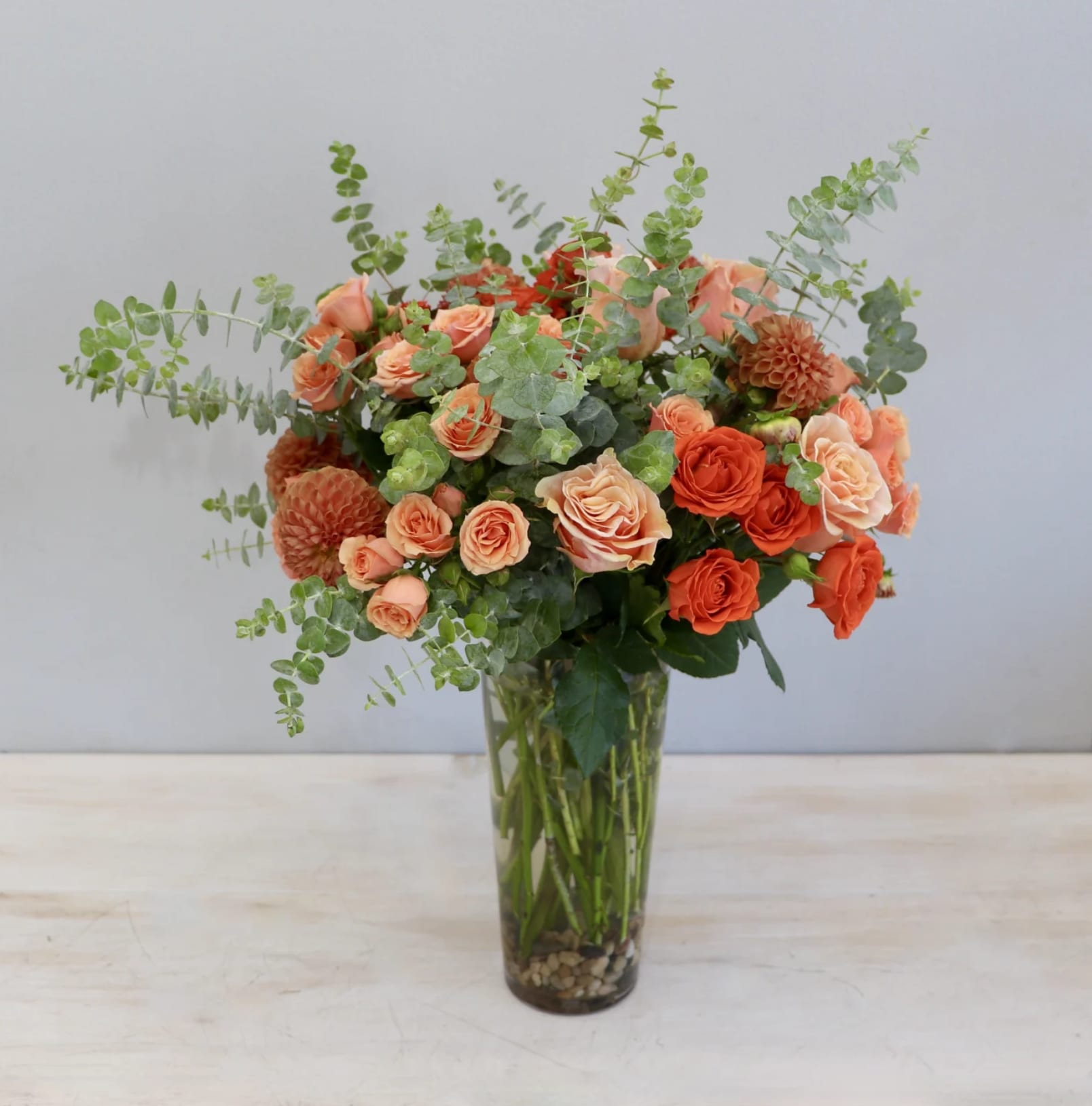 Tall glass vase of peach and orange roses with dahlias and eucalyptus accents