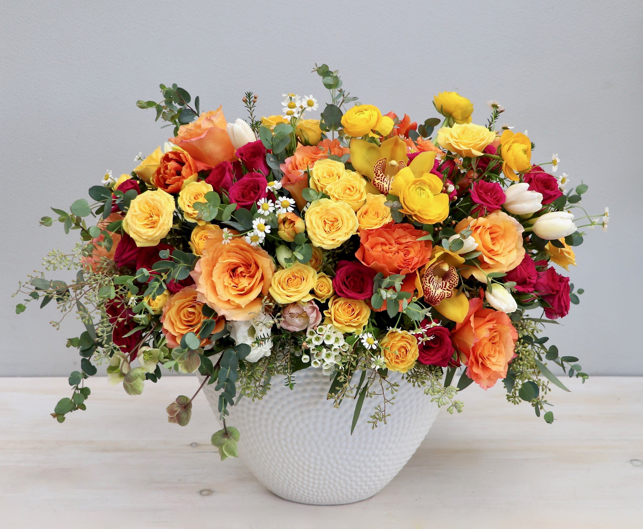 Low arrangement of yellow, orange, and red roses with ranunculus and tulips in a white ceramic bowl