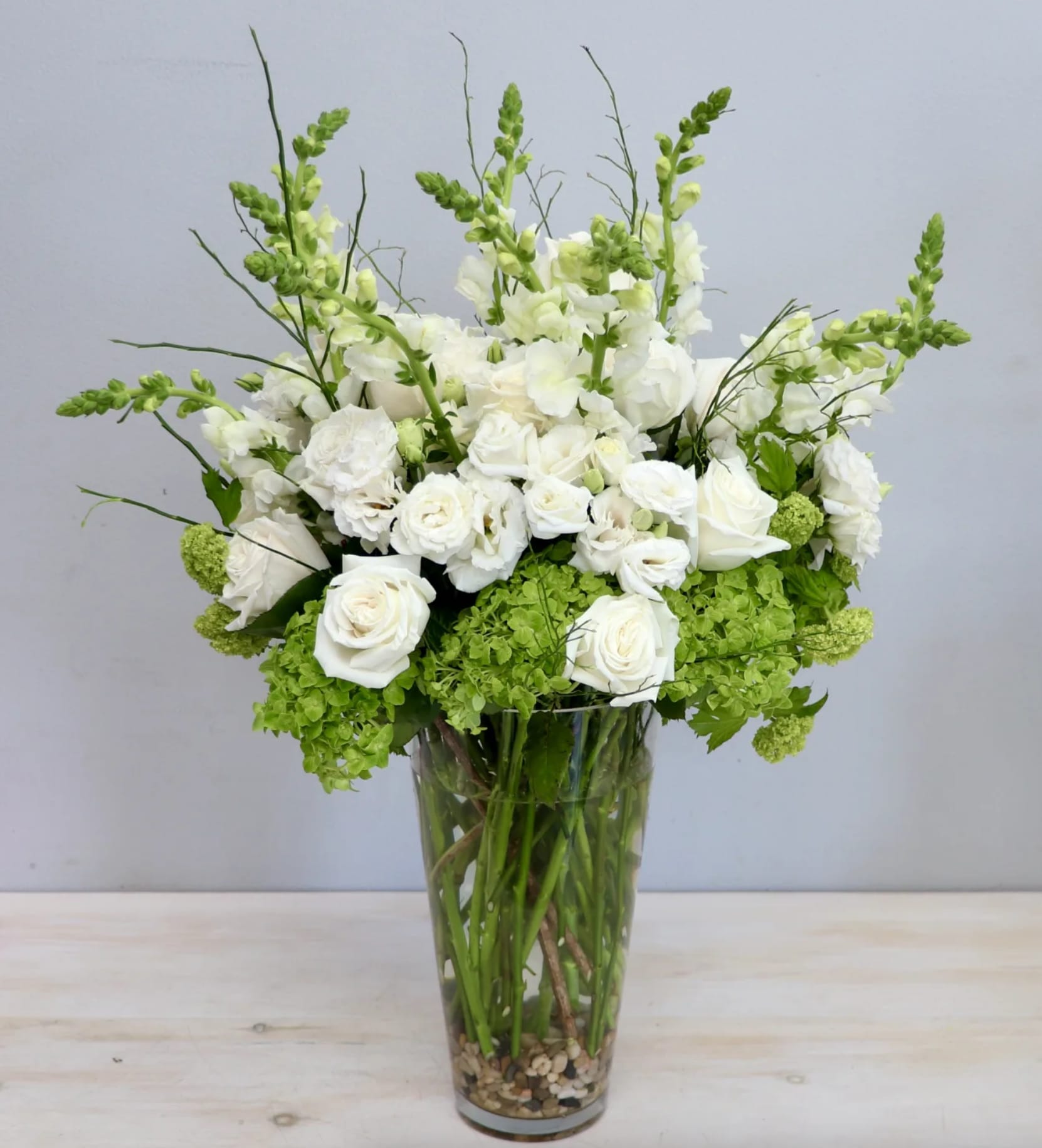 Tall arrangement of white roses and snapdragons with green hydrangeas in a clear glass vase with pebbles