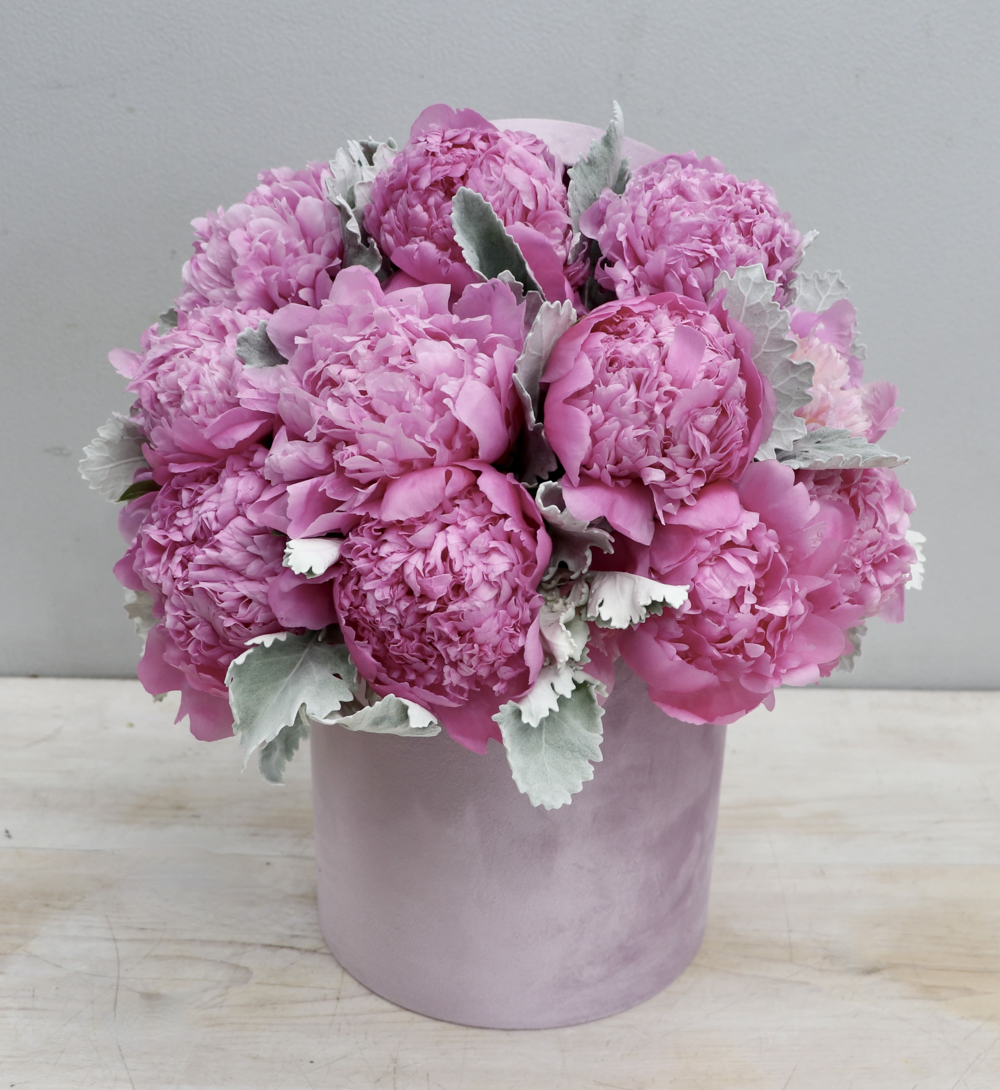 Pink peony arrangement in a round pale pink hatbox-style container with soft silvery foliage.