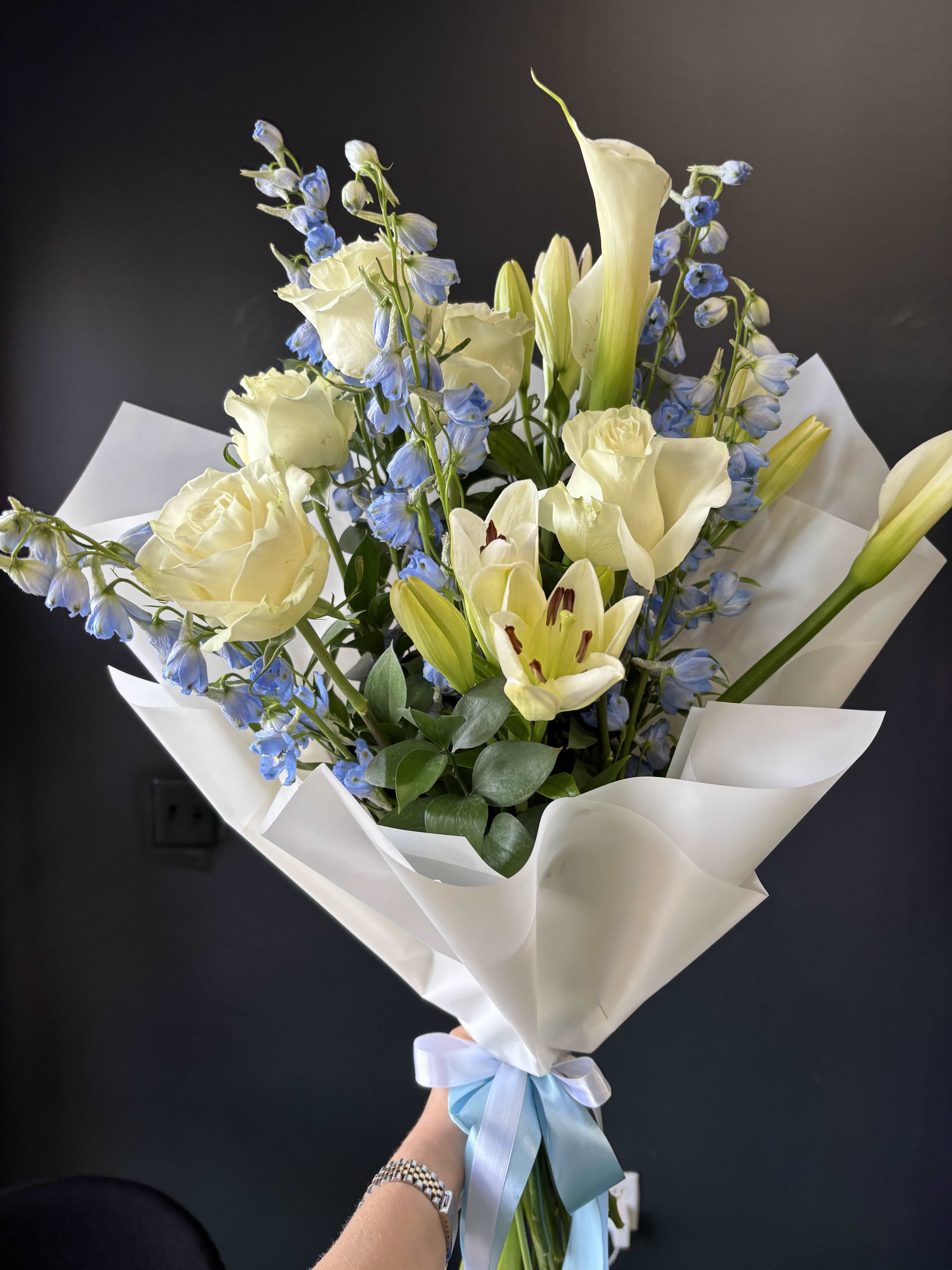 Calla Élan Bleu - A refined and airy arrangement featuring a statement ivory calla lily, creamy roses, and delicate blue delphinium, complemented by fresh greenery and soft textures. The composition blends elegance with a subtle touch of color, creating a sophisticated and modern floral design. Perfect for stylish gifts, celebrations, or moments that call for understated luxury.