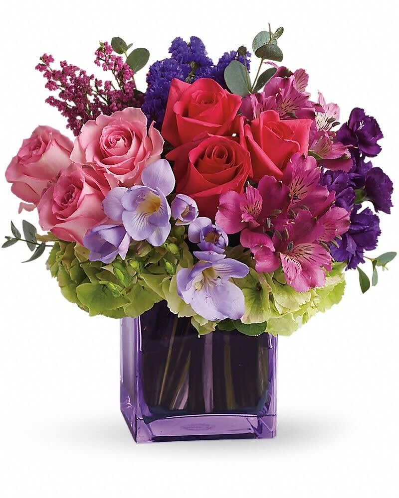 Compact arrangement of pink and red roses with purple flowers in a square purple glass vase