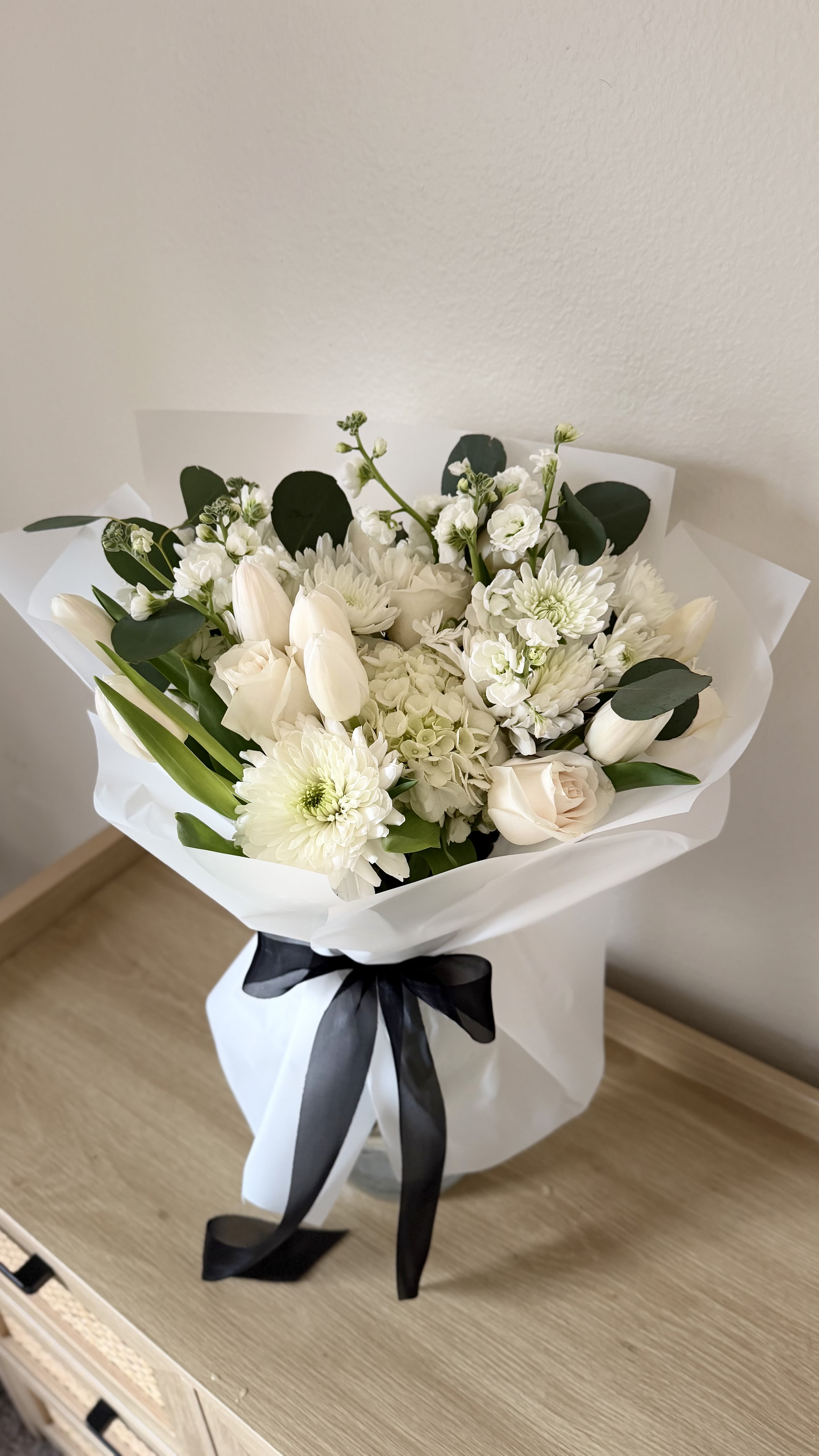 In Loving Memory - Soft, rounded clusters of white tulips, roses, chrysanthemums, hydrangeas, and stock create a serene sympathy bouquet designed to comfort and honor. This flower arrangement for memorials and sympathy gestures reads as calm and refined-the monochromatic white palette and gentle textures make it an appropriate and respectful choice when you want the person you're thinking of to feel truly remembered.  The blooms are tightly gathered into a plush, rounded shape and presented in crisp white paper wrapping tied with a wide black ribbon for a modern, minimalist contrast; a clear glass vase option is available for a lasting display. Expect a mix of velvety rose petals, sculpted tulip silhouettes, full hydrangea heads, cushiony chrysanthemums, and fragrant stock for subtle scent and visual depth. The neutral white and green tones keep the look formal and timeless.  This arrangement is suited to funerals, memorial services, or to send as a thoughtful sympathy gift that lets someone know you care. Simple care-fresh water and a cool location-will help the flowers stay dignified through services and visits; choose vase delivery when you'd like an immediate, ready-to-display tribute.