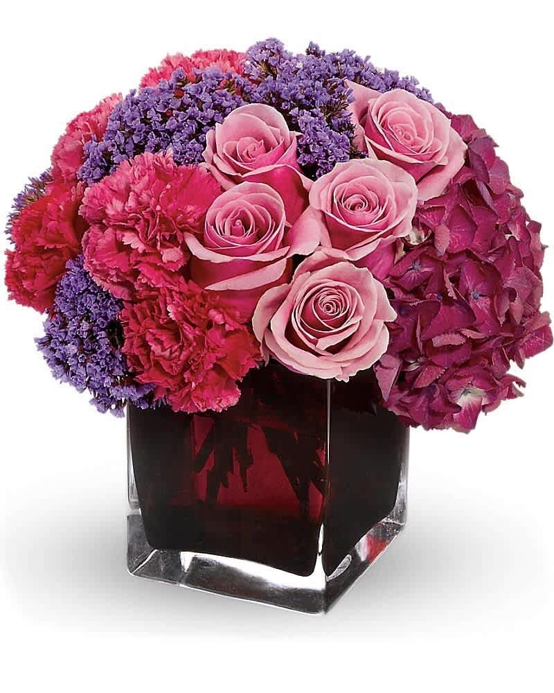 Compact arrangement of pink roses, carnations and purple flowers in a clear glass cube vase