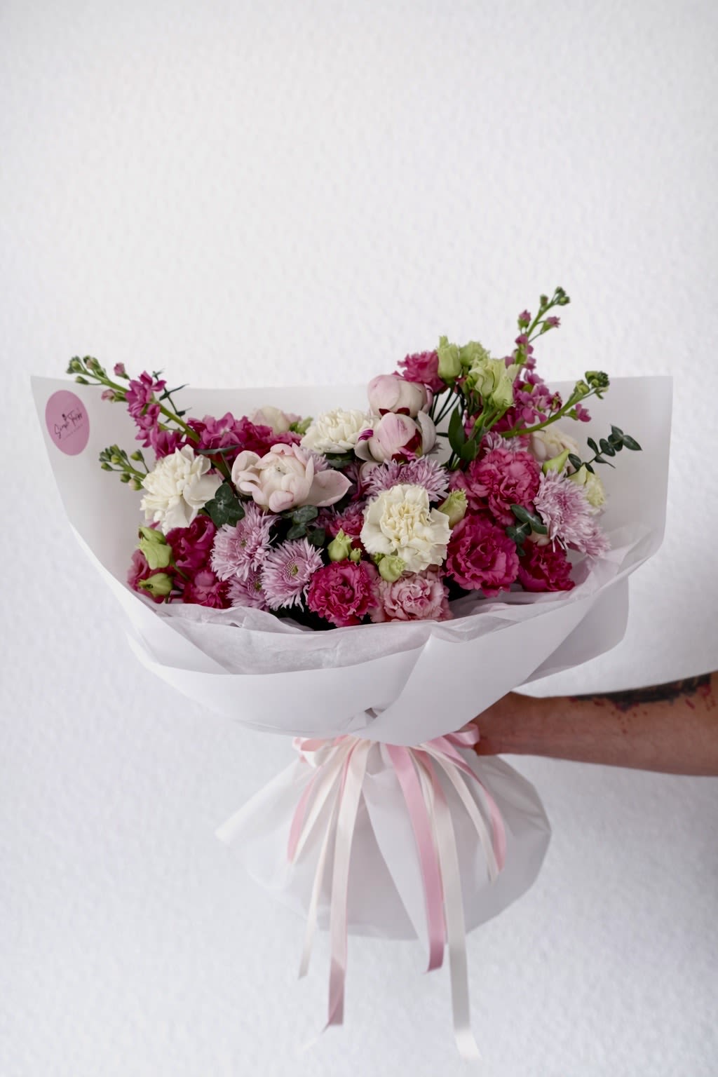 Pink bloom - Soft blush peonies and bright pink blooms mingle with creamy white carnations and pale lavender chrysanthemums in a rounded hand-tied bouquet made for birthdays, anniversaries, or any tender moment you want to mark. This flower arrangement presents a gentle, celebratory mood-perfect when you want the person you're thinking of to feel seen, loved, and comforted.  The bouquet combines peonies, carnations, chrysanthemums, lisianthus, and stems of stock for a mix of ruffled texture and airy, romantic shape. Colors range from warm pinks to creamy white with subtle lavender accents, assembled into a full, rounded silhouette and layered in crisp white paper. Finished with cascading pink and white satin ribbons and a round brand sticker, the presentation arrives gift-ready and refined.  Ideal for Mother's Day, a new baby visit, get-well wishes, romantic gestures, or a graceful sympathy offering, this arrangement is designed to convey affection and gentle support. Hand-tied for easy display, refresh the water daily and trim stems if transferring to a vase to extend the life of the flowers.