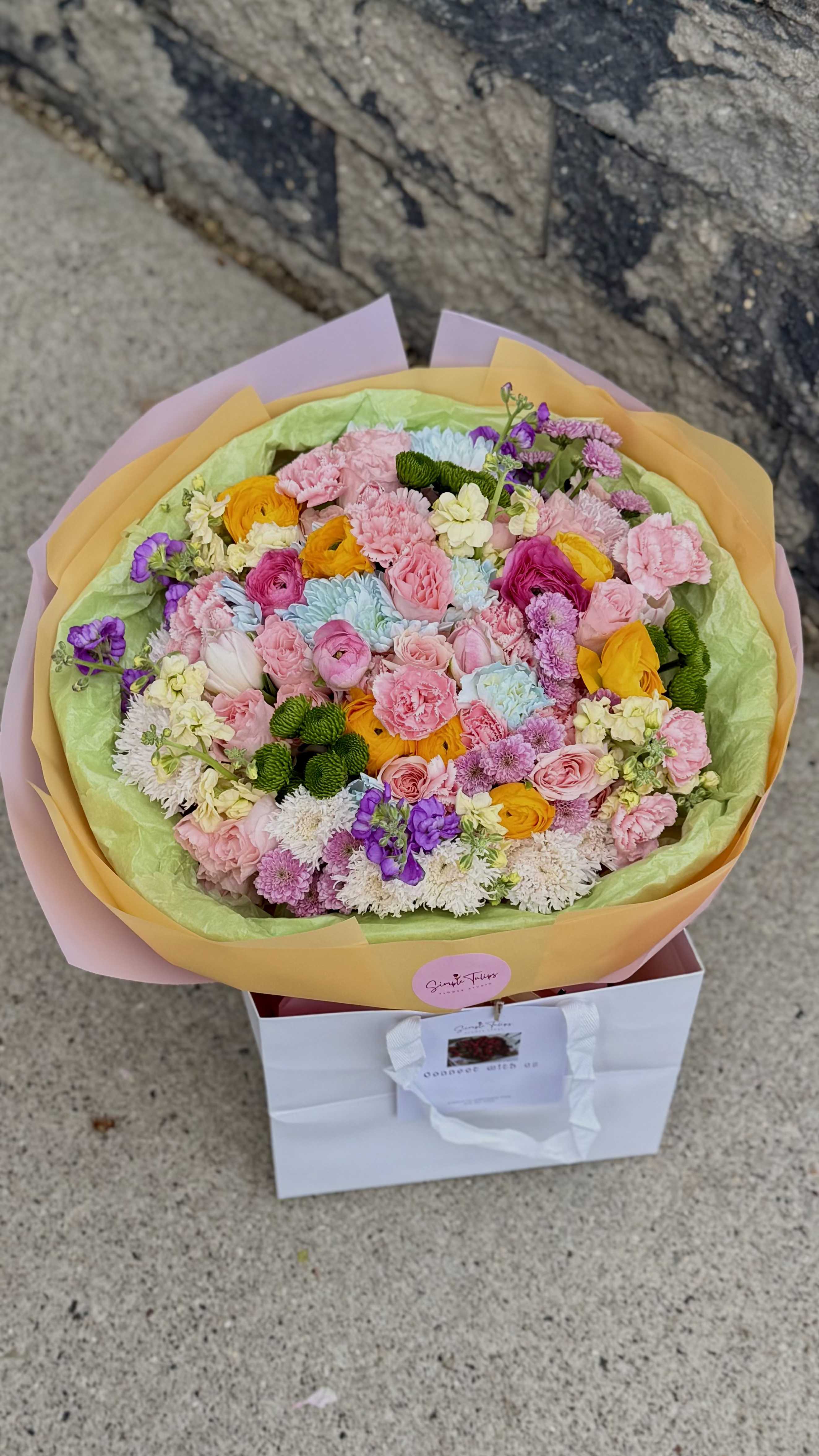 Huge dopamine bouquet - A lush, round cluster of pastel blooms designed to spark joy at birthdays, Mother's Day, new baby celebrations, or any moment that calls for a cheerful surprise. This flower arrangement places the recipient front and center-she'll feel celebrated the moment the gift bag is opened and the carpet-like surface of pink roses and carnations greets her with bright, optimistic color.  The bouquet is packed tight in a low, round box-style container wrapped in layers of soft pastel tissue paper for an elegant, finished look. Pink dominates the palette, balanced by yellow and orange ranunculus, lavender stock, white button chrysanthemums, and pale-blue accents that create lively contrast and texture. The arrangement sits inside a white paper shopping bag with handles and a branded hang tag, so it arrives ready to present.  Perfect for the person you want to brighten-whether you're saying happy birthday, congratulations, or simply thinking of them-this boxed bouquet is easy to display on a table or counter. Size and care details are provided at checkout so you can choose the right scale and keep the blooms fresh after delivery.