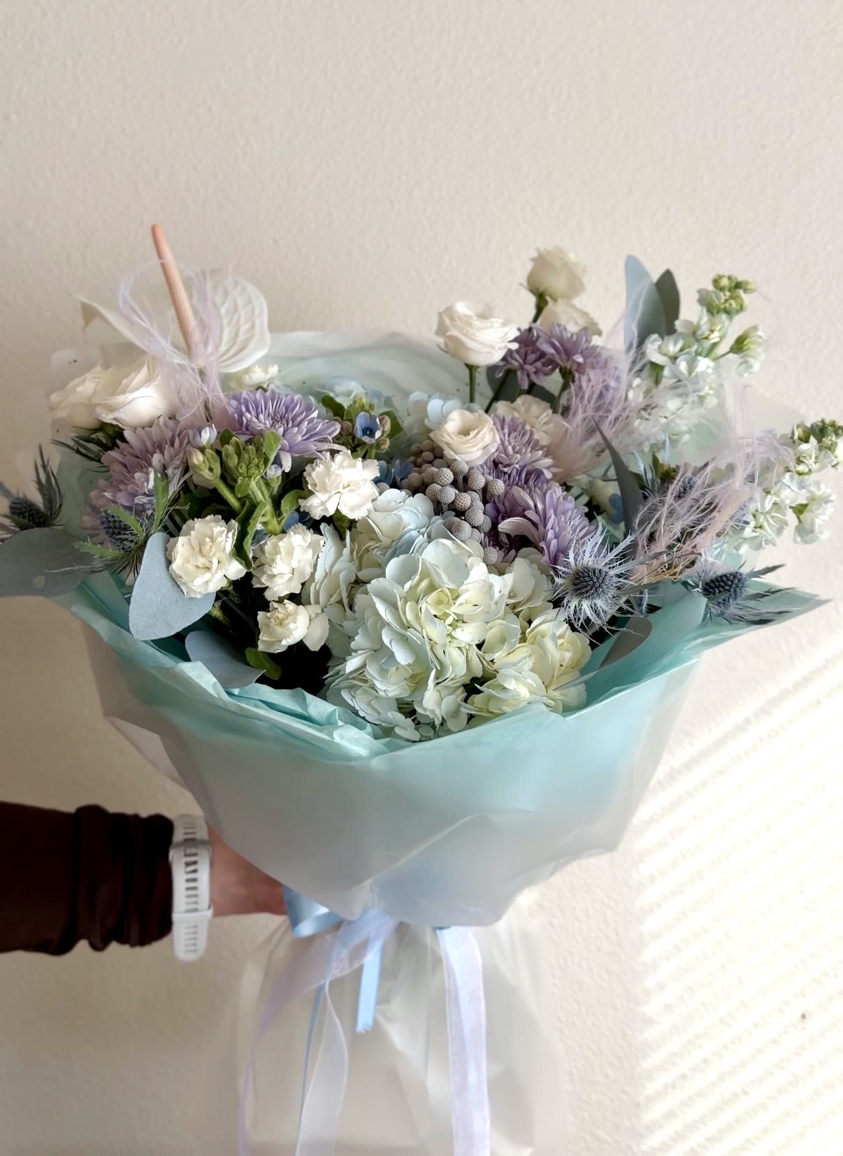 Frozen Bouquet - A cool, airy mix of white and lavender flowers makes this hand-tied bouquet a serene choice for a birthday bouquet, a get-well gift, or simply to let someone know you're thinking of them. The Frozen Bouquet's soft color story and modern accents give the recipient an immediate sense of calm and thoughtful care-perfect for marking celebrations or offering comfort without being overwhelming.  Large white hydrangeas anchor the front of the arrangement, while clusters of small white carnations and spray roses nestle among lavender chrysanthemums and tall, fragrant stock for layered texture. Thistle-like accents, delicate berries, an anthurium-style bloom, and wispy pastel feathers lend a contemporary, airy feel. The bouquet is wrapped in translucent white and aqua-blue cellophane with coordinating tissue and ribbon for a finished, gift-ready look.  This arrangement suits someone who appreciates cool, sophisticated palettes-friends, family, or colleagues who deserve a gentle, elegant surprise. Hand-tied for an organic shape, it arrives ready to place in water; standard care-fresh water, a clean vase, and a fresh cut to stems-will keep the blooms looking their best.