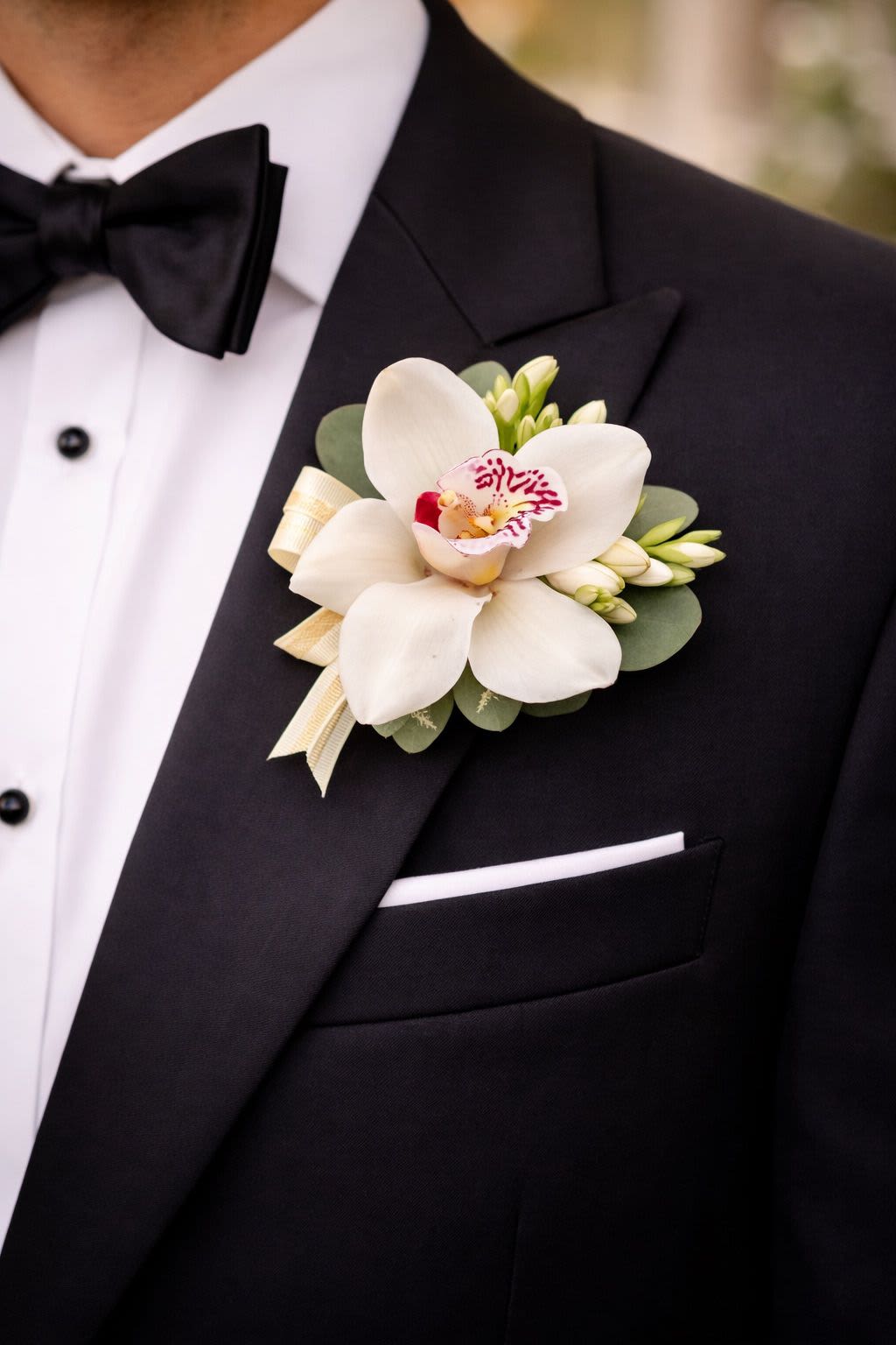 Boutonniere Orchid by The House of Flowers - A refined and elegant boutonniere featuring a fresh orchid bloom, accented with delicate greenery and soft complementary buds. Carefully handcrafted with a clean and sophisticated finish, this piece is perfect for weddings, proms, and special occasions.  Available colors: white, red, green, and yellow. Custom colors are available upon request with advance notice. Elegantly packaged in a beautiful presentation box. 