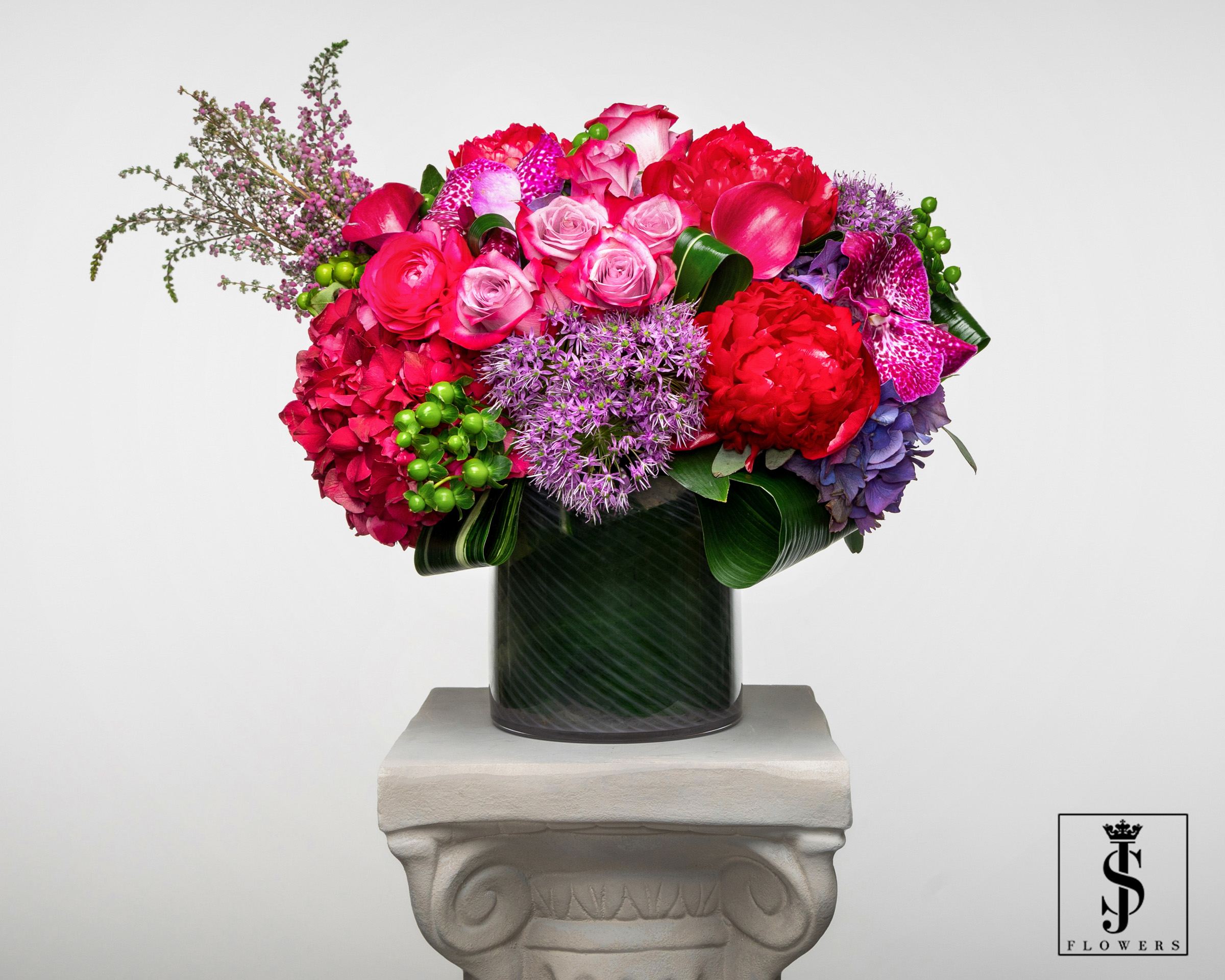 The Jeweled Romance  -  Introducing our exquisite floral creation: Jeweled Romance!  Let the deep tones of burgundy hydrangea and luscious purple lipstick roses ignite your passion.  The vibrant touch of green hypericum  berries adds a playful element, while the delicate pink bold peonies bring an air of romance.  The purple orchids and green aspidistra leaf add an elegant finishing touch, making this arrangement a true gem. 