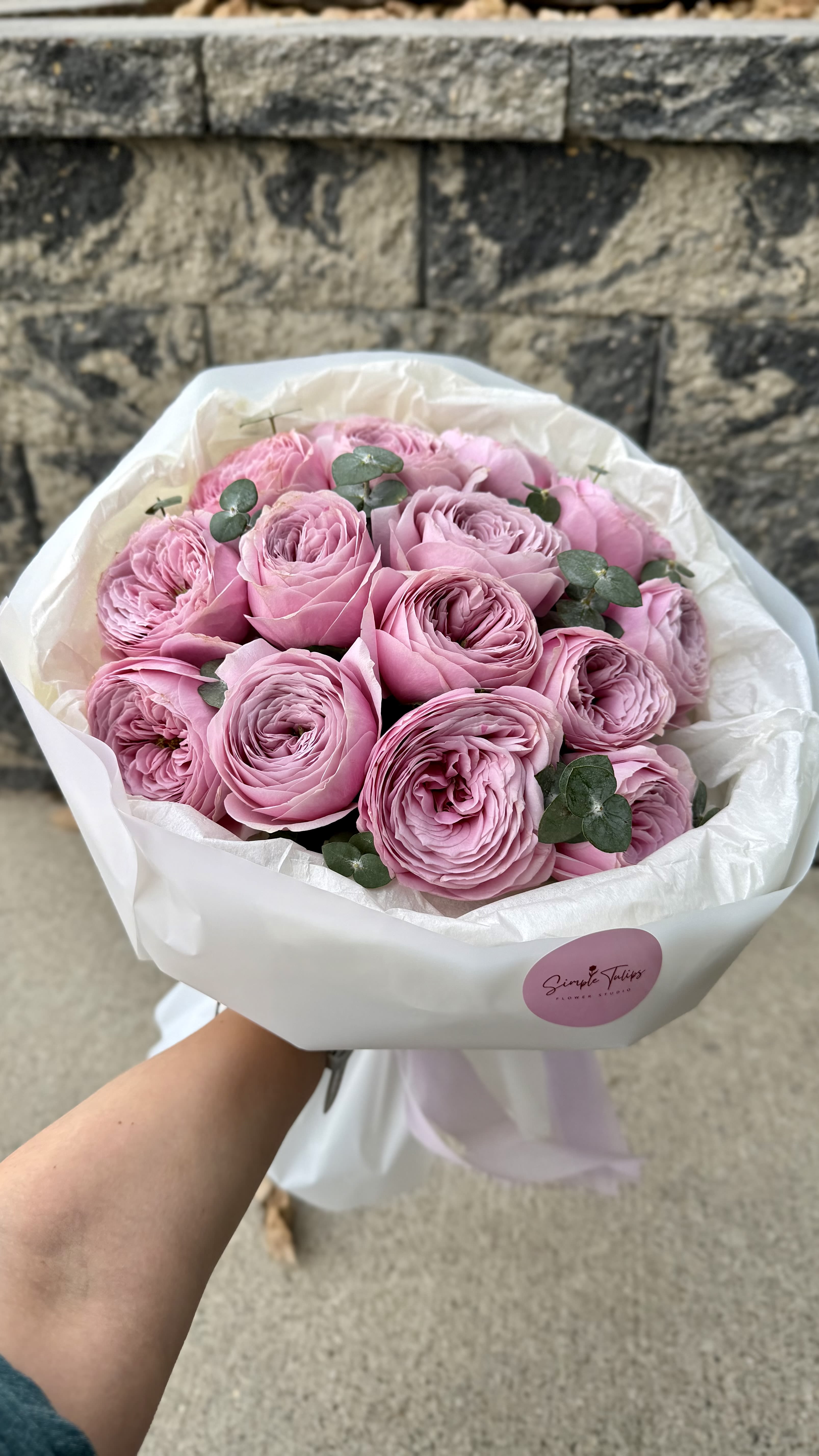 Lavender Garden Roses Bouquet  - Soft lavender garden roses arranged in a tight, rosette-packed bouquet-perfect as an anniversary bouquet or a romantic wedding gift that says "I cherish you." The compact, rounded shape and layered petals give a lush, vintage feel that will make the person you're thinking of feel truly seen and adored.  Each arrangement features pink and white garden-style roses wrapped in crisp white paper and tissue, finished with a soft ribbon and brand sticker for a polished presentation. The dense cluster highlights the roses' full, velvety texture and gentle color palette, creating an elegant, tactile impression that holds up well on a dining table or bedside.  Ideal for anniversaries, weddings, or heartfelt celebrations, this bouquet arrives ready to display-no vase needed for the initial impression. To keep the roses looking fresh, trim stems and place in clean water with floral food when you're ready to display; sizing and exact stem count vary with seasonal availability.
