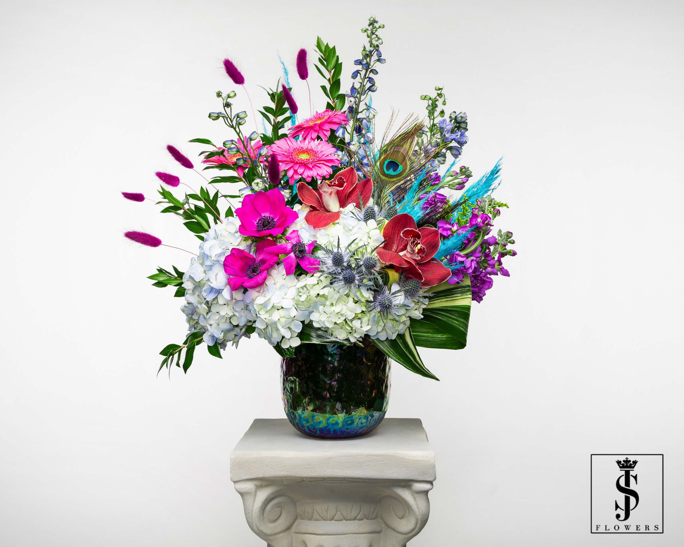 Tall mixed arrangement with white hydrangeas, bright pink blooms, orchids and peacock feather in iridescent vase.