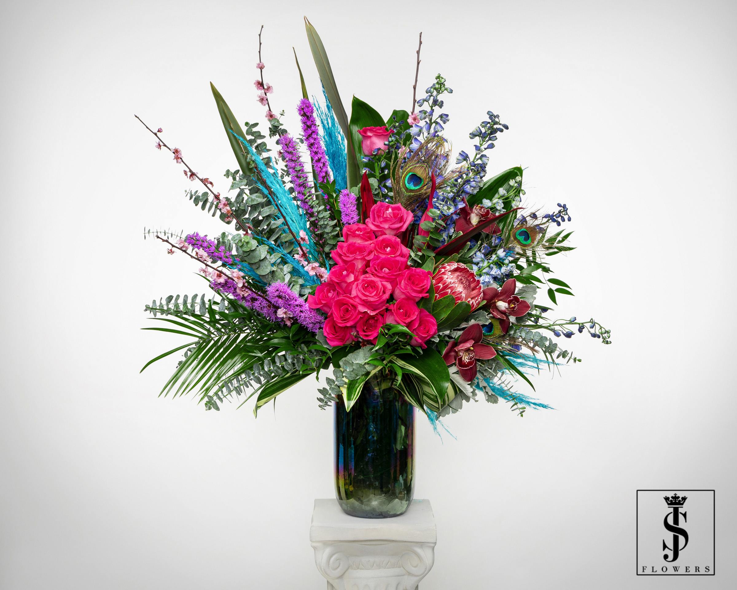 Tall arrangement of hot pink roses, orchids, protea, and peacock feathers in a dark glass vase