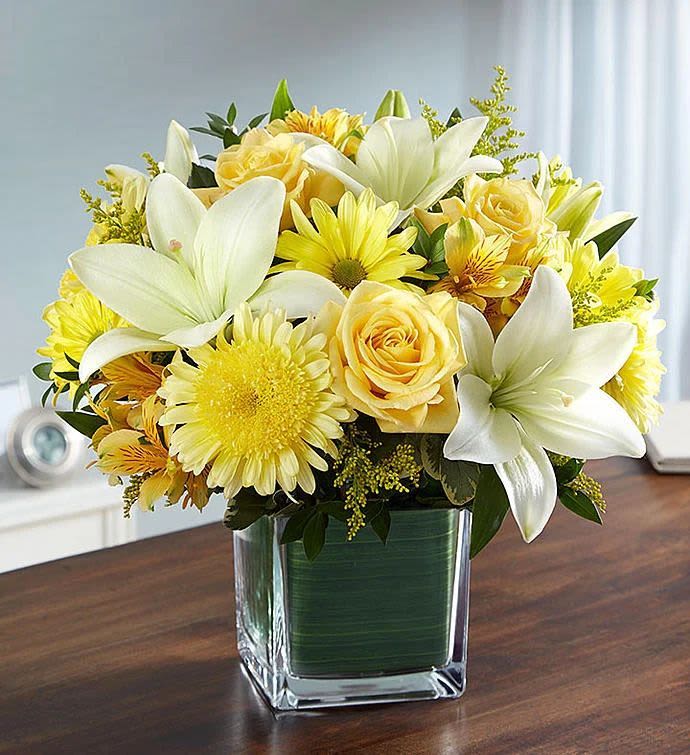 Golden Harmony Arrangement - Pale yellow roses and large white lilies form a sunny, uplifting centerpiece-perfect as a cheerful Mother's Day bouquet, a get-well gift, or a just-because surprise. This compact flower arrangement brings warm yellow daisies and soft white blooms together in a tidy, low-profile design that reads fresh and optimistic for birthdays, graduations, anniversaries, or to brighten an office desk.  Set in a clear glass cube vase, the arrangement features pale yellow roses, bright yellow daisy-style flowers, and prominent white lilies, with broad green leaves lining the inside of the container as a visible design accent. Its small footprint makes it ideal for side tables, workspaces, or intimate centerpieces; the clean cube keeps the composition neat while showcasing stems and foliage. Order with confidence for occasions from thinking-of-you to sympathy or housewarming-this easy-care, compact display arrives ready to place and enjoy.