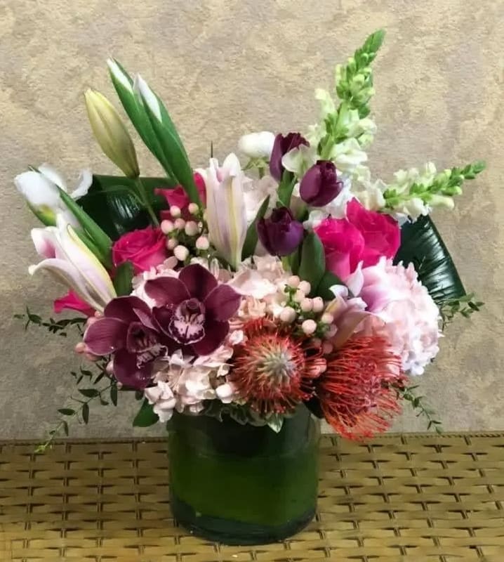 Porto, Portugal - Recipe: White Snapdragons, Pink Hydrangeas, Purple Cymbidium Orchids, Stargazer Lilies, Pink Peonies (Seasonal), White Ranunculus (seasonal), Hot Pink Roses, Purple Tulips, White Iris, Orange Pincushion, Pink Hypericum Berries, and Leaves in a short vase with leaf wrap. Availability: Peonies November-mid-August Substitute Available: Yes Design View: All Facing View Photo shown: Regular 