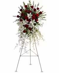 Lily and Rose Tribute - Premium - A tall, graceful standing spray that blends deep red roses, large white lilies, and delicate orchids into a dignified tribute - ideal for sympathy and funeral services where a respectful, memorable statement is needed. The elongated, cascading silhouette tapers gently, creating a flowing profile that draws the eye downward and conveys quiet reverence for the person you're honoring.  The arrangement features a dense central cluster of velvety red roses set against the sculptural white lilies, with orchids woven throughout for a refined, textured contrast. Fine-textured filler and trailing green stems soften the edges and add subtle movement, while fresh greenery provides a rich backdrop that lets each bloom stand out. Mounted on a slim metal easel stand for upright display, this piece is sized to command presence at a chapel, memorial venue, or graveside.  Choose this tribute when you want the recipient to feel genuinely remembered and comforted-its formal lines and timeless palette speak of love and respect without words. Please include service details and delivery location with your order so we can ensure proper placement on the easel at the venue.