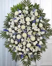 Blue Horizon Remembrance Spray - A graceful and elegant blue and white standing sympathy spray designed to express peace, comfort, and remembrance. This beautiful tribute features white roses, blue delphinium, fragrant stock, white cushion mums, and white cremon mums, thoughtfully arranged and filled with delicate white monte casino asters and lush fresh greenery.  The calming combination of blue and white flowers symbolizes peace, honor, and everlasting love, creating a dignified display of sympathy. Presented on a standing easel, this arrangement offers a heartfelt way to honor a cherished life and provide comfort during a time of loss. 