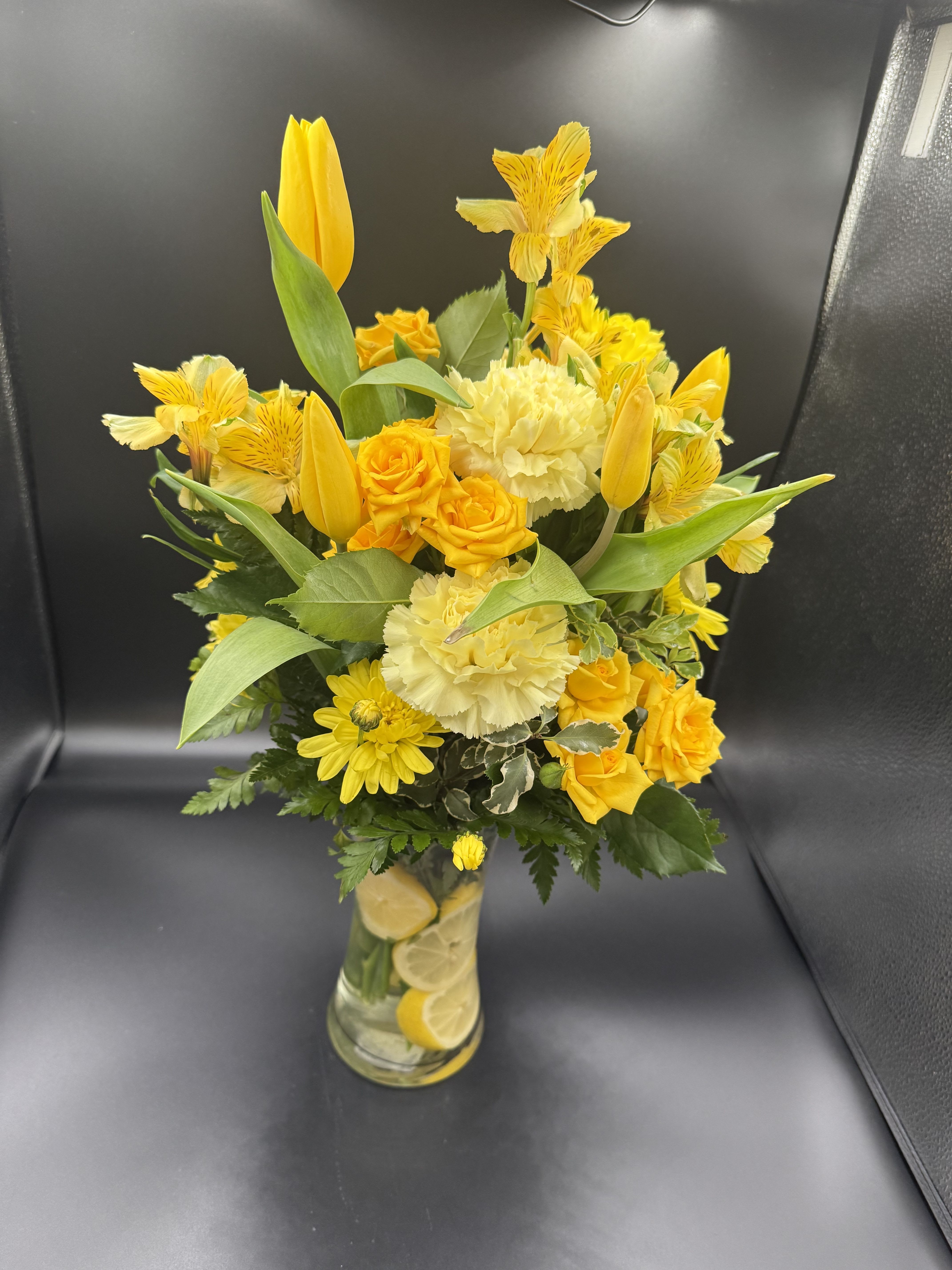 Tall yellow bouquet with tulips, roses, carnations and daisies in a clear vase with lemon slices