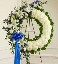 Serene White &amp; Blue Tribute Wreath - A graceful and elegant solid white wreath, thoughtfully designed to convey peace and remembrance. This arrangement features a lush base of pristine white blooms, including soft white carnations, elegant white lilies, and delicate white spray roses. Accented with striking blue delphinium, the design offers a subtle touch of color symbolizing tranquility and honor. Finished with a beautifully coordinated blue bow, this wreath creates a heartfelt tribute, perfect for expressing sympathy and everlasting love.