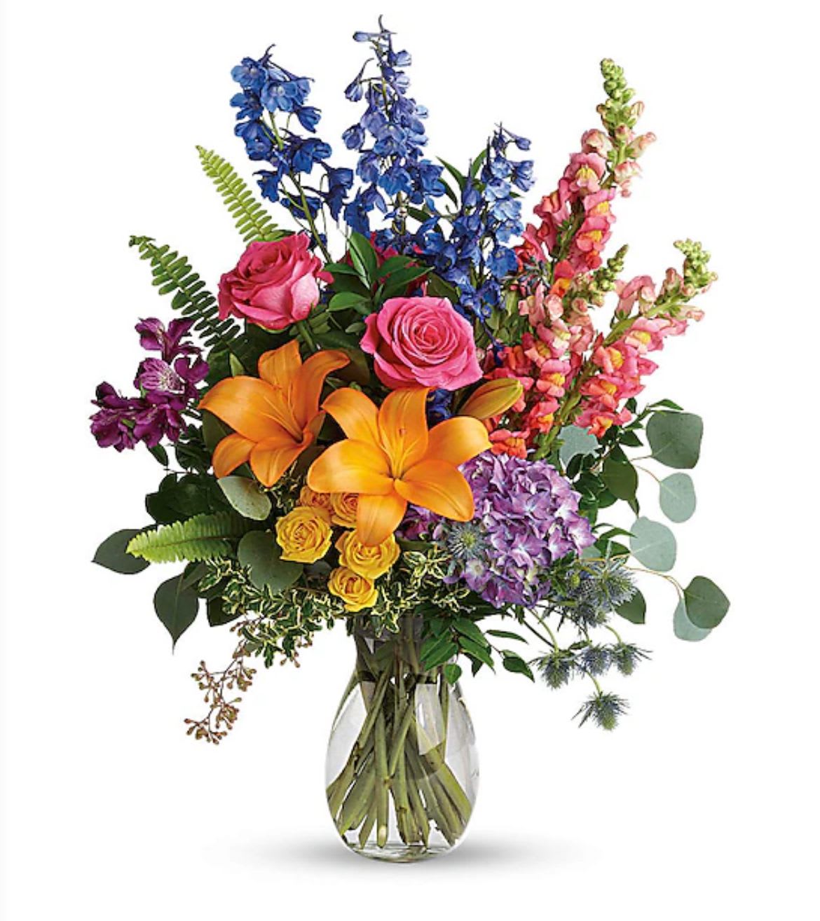 Tall mixed bouquet with orange lilies, pink roses, blue delphinium and purple hydrangea in a clear glass vase