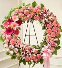 Blushing Remembrance Standing Wreath - A beautiful pink standing wreath designed to honor and celebrate a cherished life. This elegant tribute features a graceful blend of pink carnations, pink gerbera daisies, roses, spray roses, and oriental stargazer lilies, thoughtfully arranged with fresh greenery for a full and balanced display.  Finished with a pink ribbon bow, this wreath symbolizes love, compassion, and remembrance, offering a heartfelt and dignified expression of sympathy for funeral or memorial services.