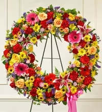 Vibrant Garden Tribute Standing Wreath - A bold and cheerful standing wreath bursting with vibrant colors and textures. This lively tribute features yellow daisies, orange gerbera daisies, hot pink gerbera daisies, oriental stargazer lilies, red roses, red carnations, yellow roses, purple statice, and green button mums, arranged with lush fresh greenery for a full and dynamic display.  Finished with a hot pink ribbon bow, this wreath conveys love, celebration, and heartfelt remembrance, making it a striking and uplifting tribute for funeral or memorial service
