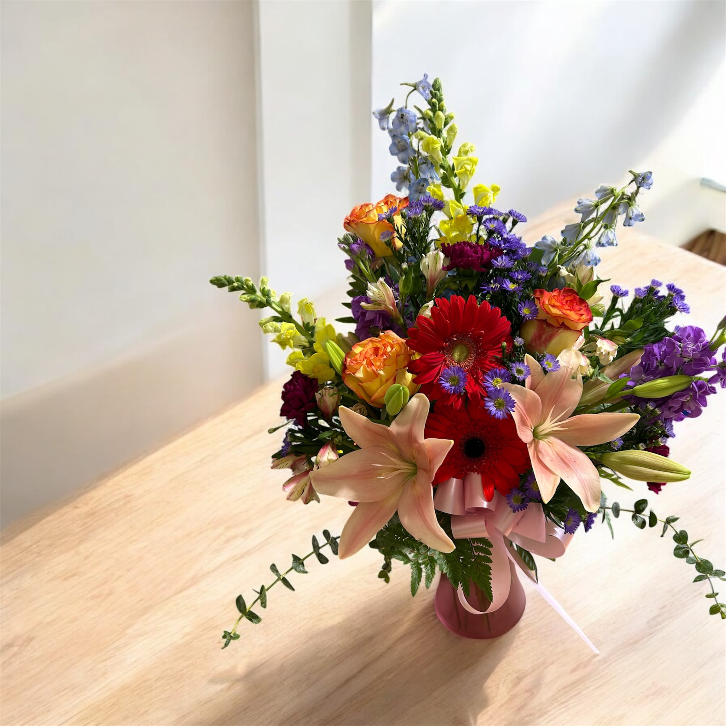 Bright mixed bouquet with lilies, roses, gerbera daisies and delphinium in a pink vase on a wooden table