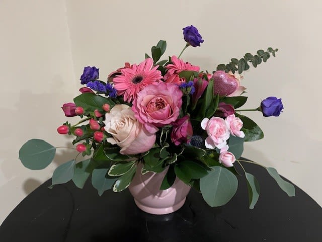 Pink and purple flower arrangement with roses and gerbera daisies in a pale pink ceramic bowl.