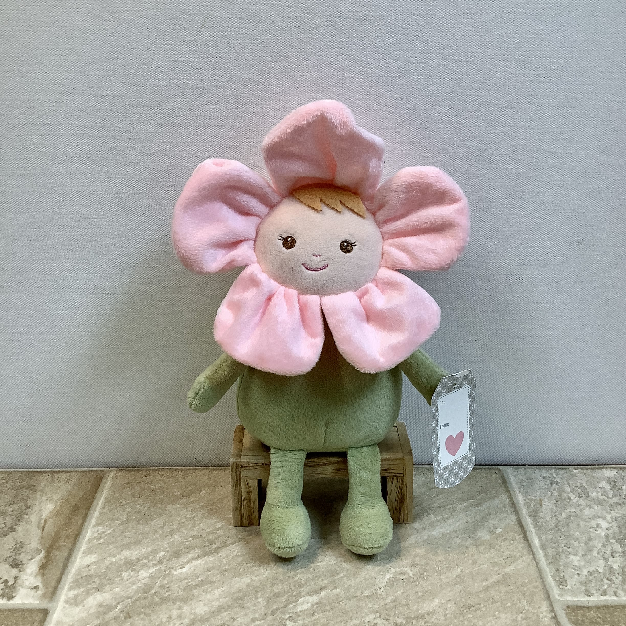 Plush flower doll with pink petals and a green body sitting on a small wooden stool