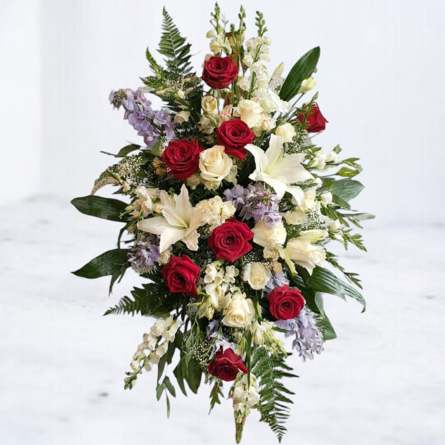 Tall standing spray of red and white roses, white lilies, and soft lavender blooms framed by ferns.
