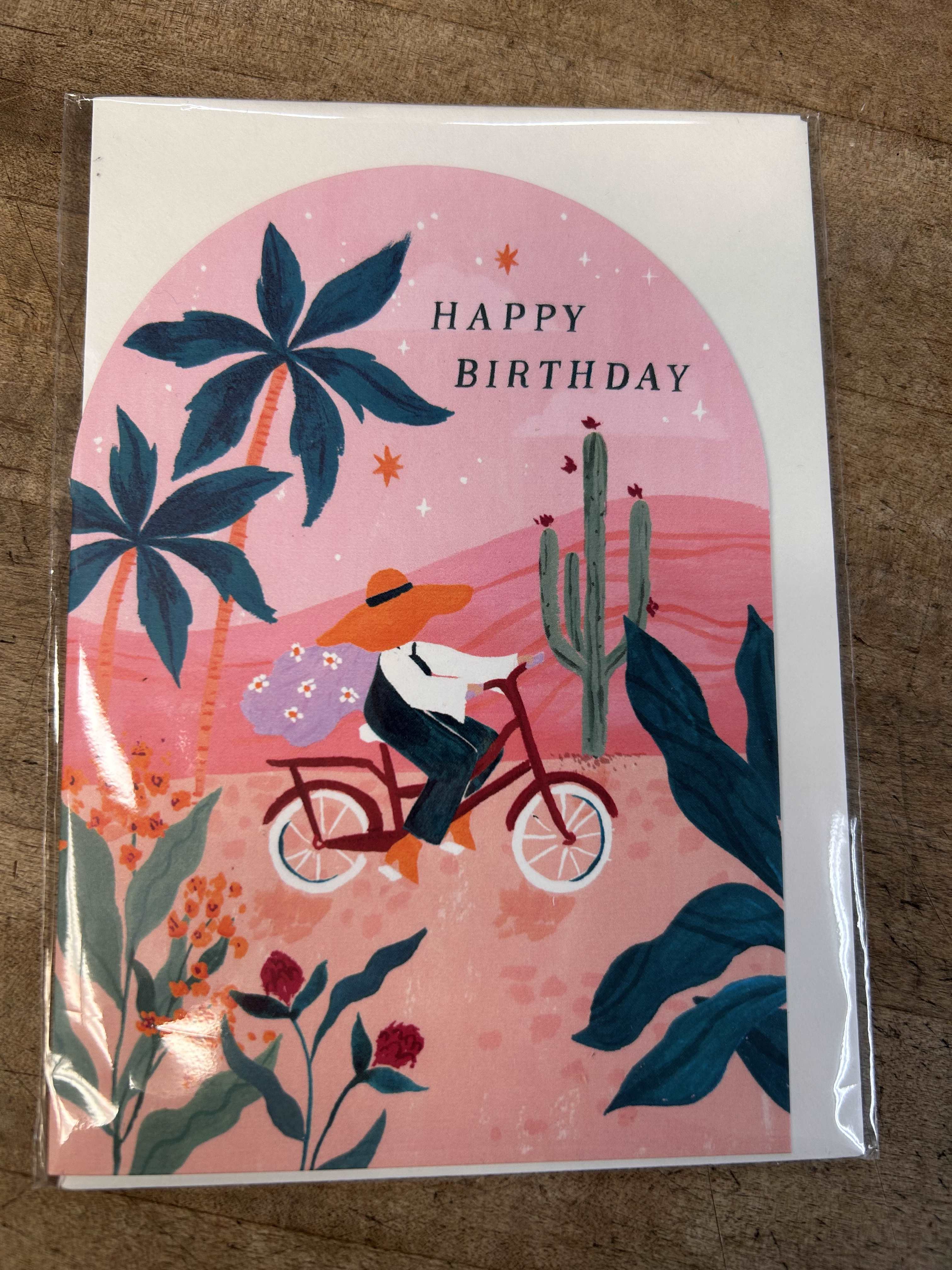 Illustrated Happy Birthday card with a person riding a bicycle through a pink desert scene