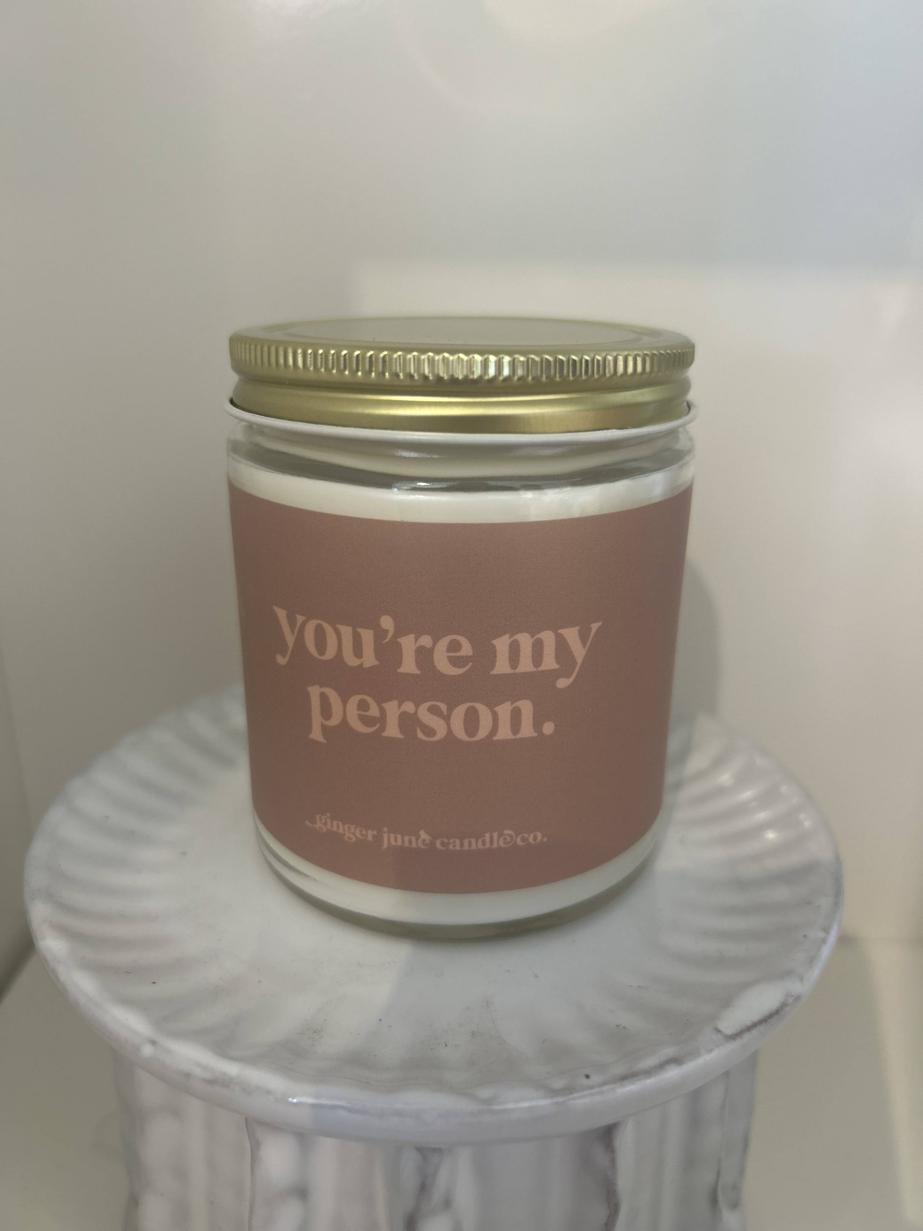 Glass jar candle with gold lid and label reading "you’re my person" on a white pedestal stand.
