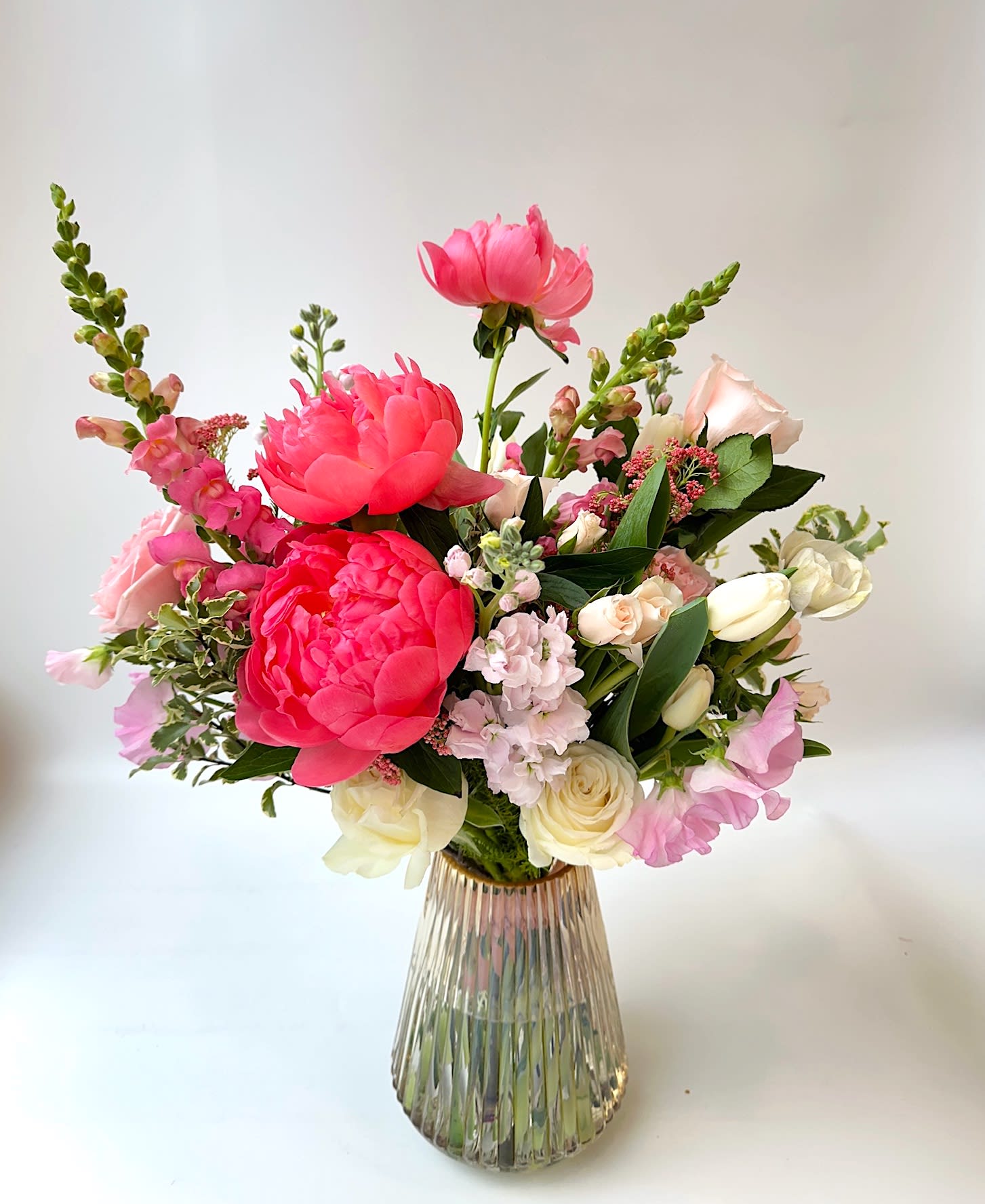 Vase arrangement of bright pink peonies with pale roses, stock, and snapdragons in a ribbed glass vase