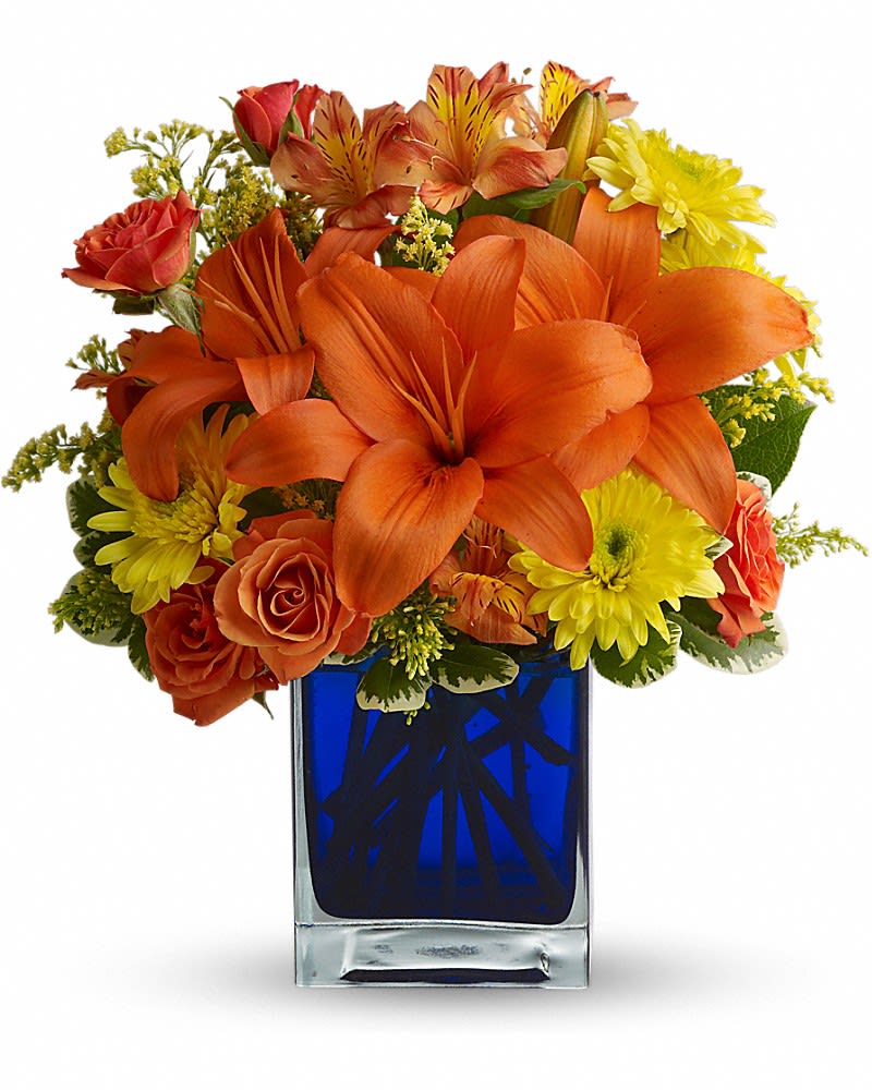 Compact arrangement of orange lilies, roses, and yellow mums in a blue glass cube vase