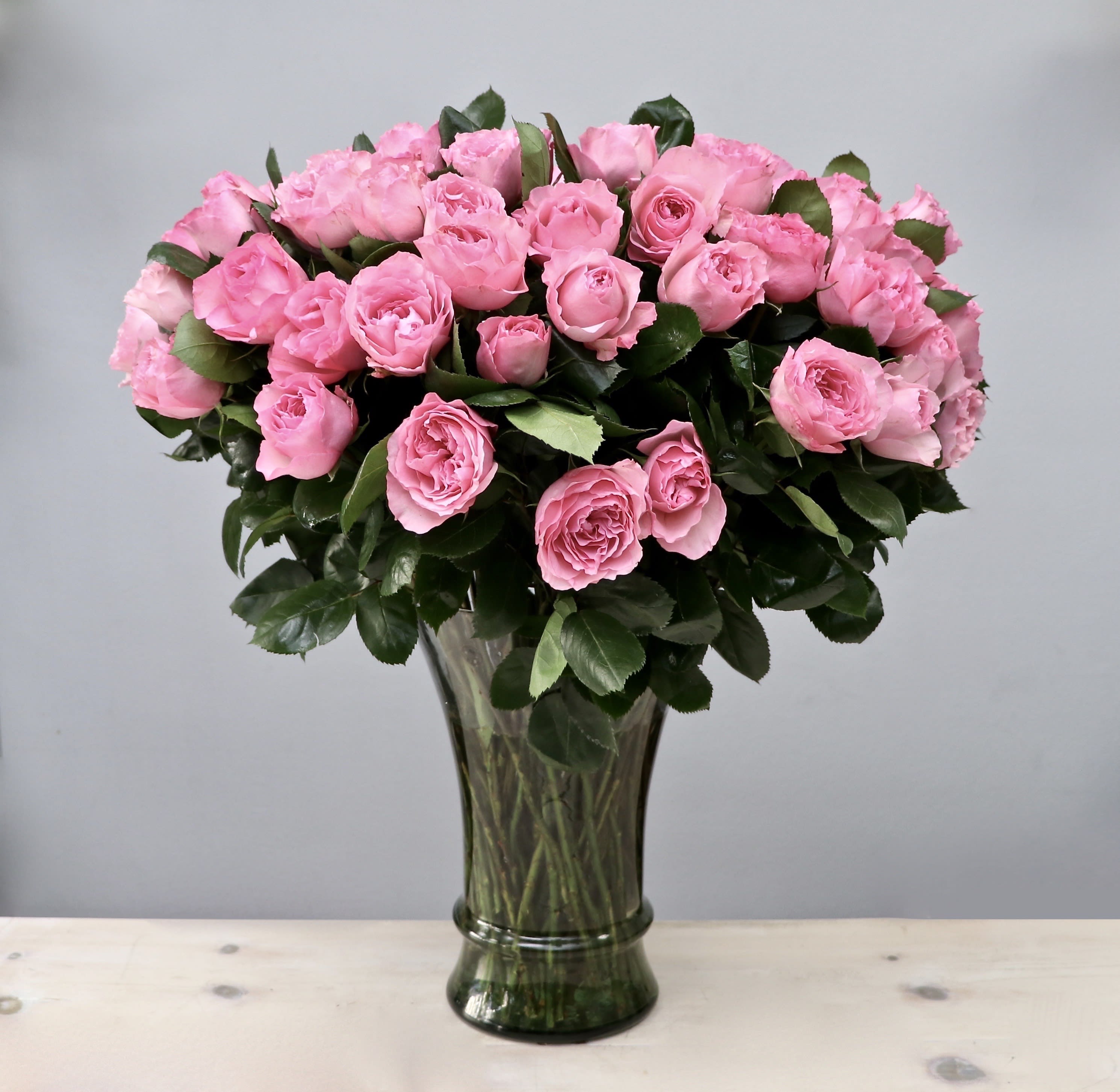 Large arrangement of pink roses in a tall clear glass vase against a gray background