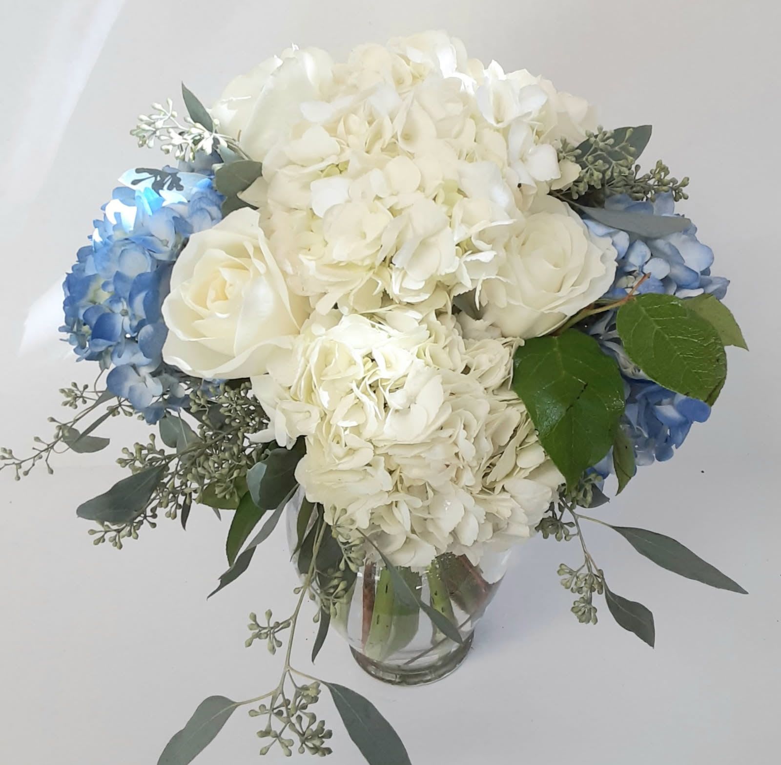 Blue and White Abundance - Ever popular Blue and white hydrangeas billow out like clouds, surrounded by white lovely roses and seeded eucalyptus in a clear vase, for any occasion. 