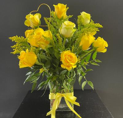 Tall arrangement of yellow roses in a clear glass vase with a yellow bow and heart-shaped accent