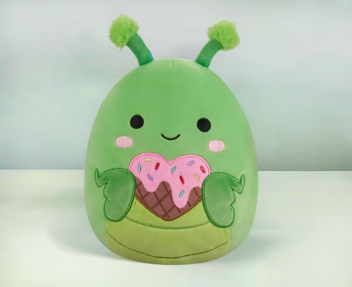 Green plush character with antennae holding a pink sprinkled heart-shaped treat.