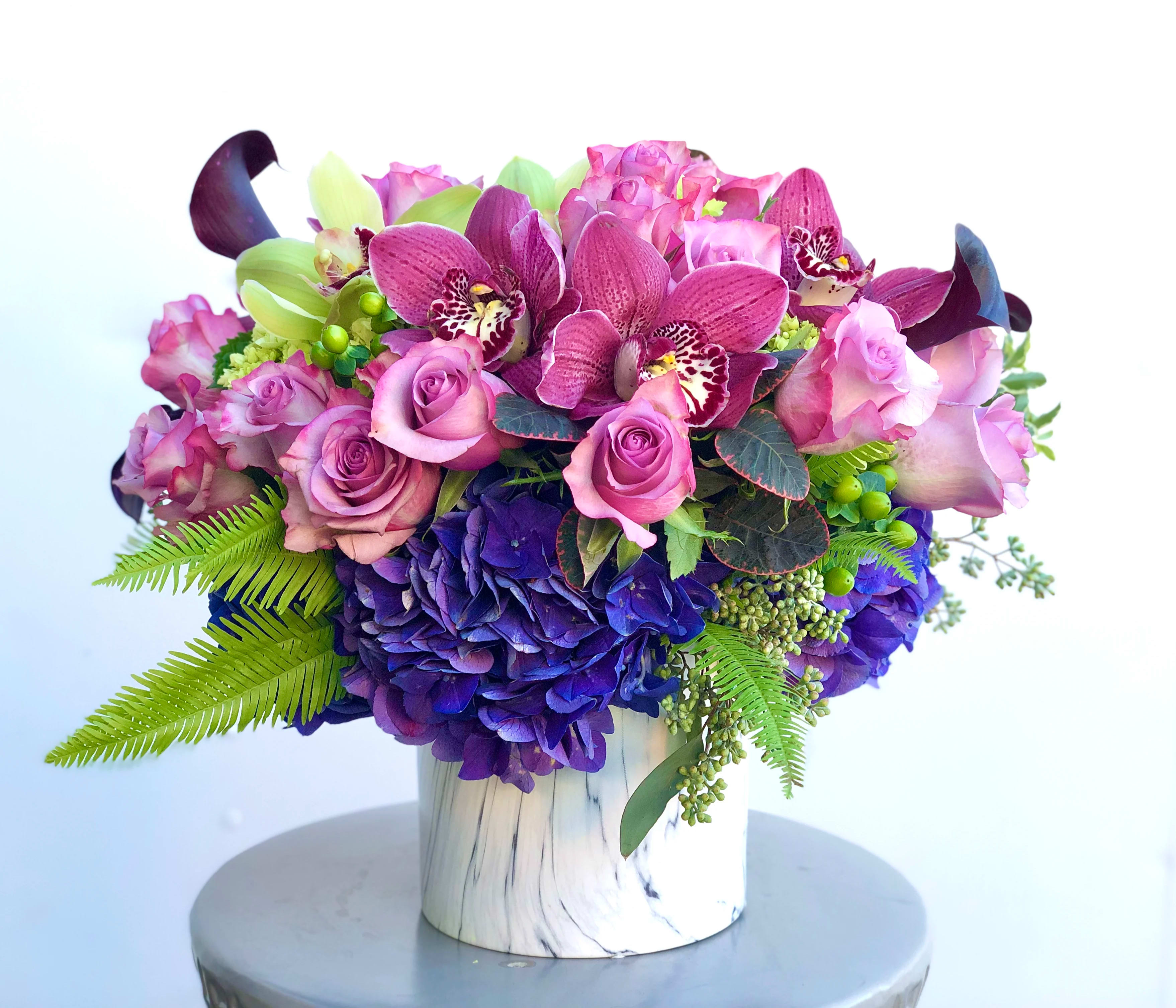 Royal Garden - This gorgeous jewel-colored arrangement is full of different shades of purple and lavender. This bright and lush piece is like a small garden in a perfectly portable ceramic vase. Roses, Hydrangea, and Calla Lilies are topped with Cymbidium Orchids