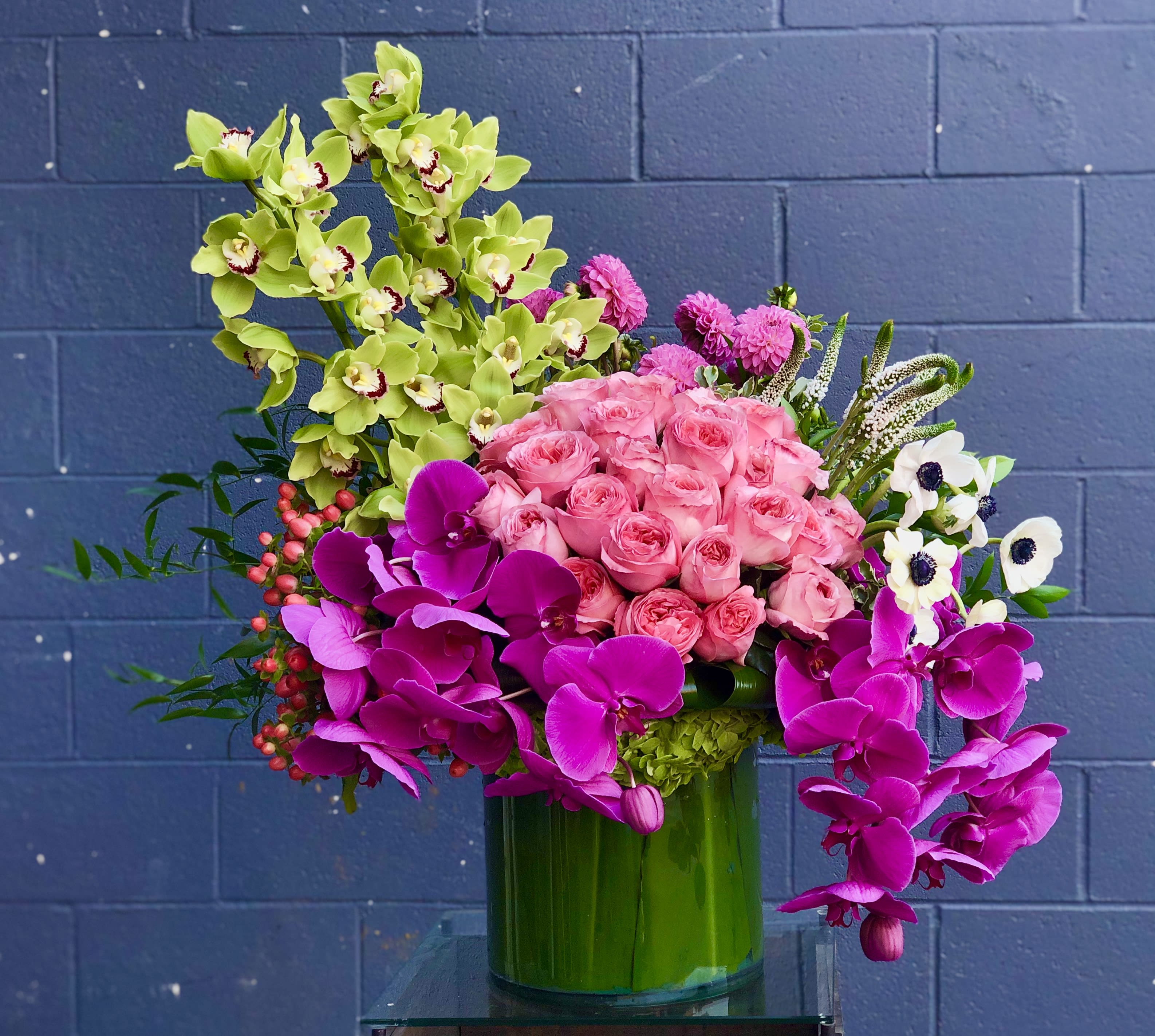 Funky Fuchsia - A bright and colorful arrangement of stunning Cymbidium and Phalaenopsis Orchids, Roses, and various other luxurious blooms in a large, leaf-lined cylinder vase.