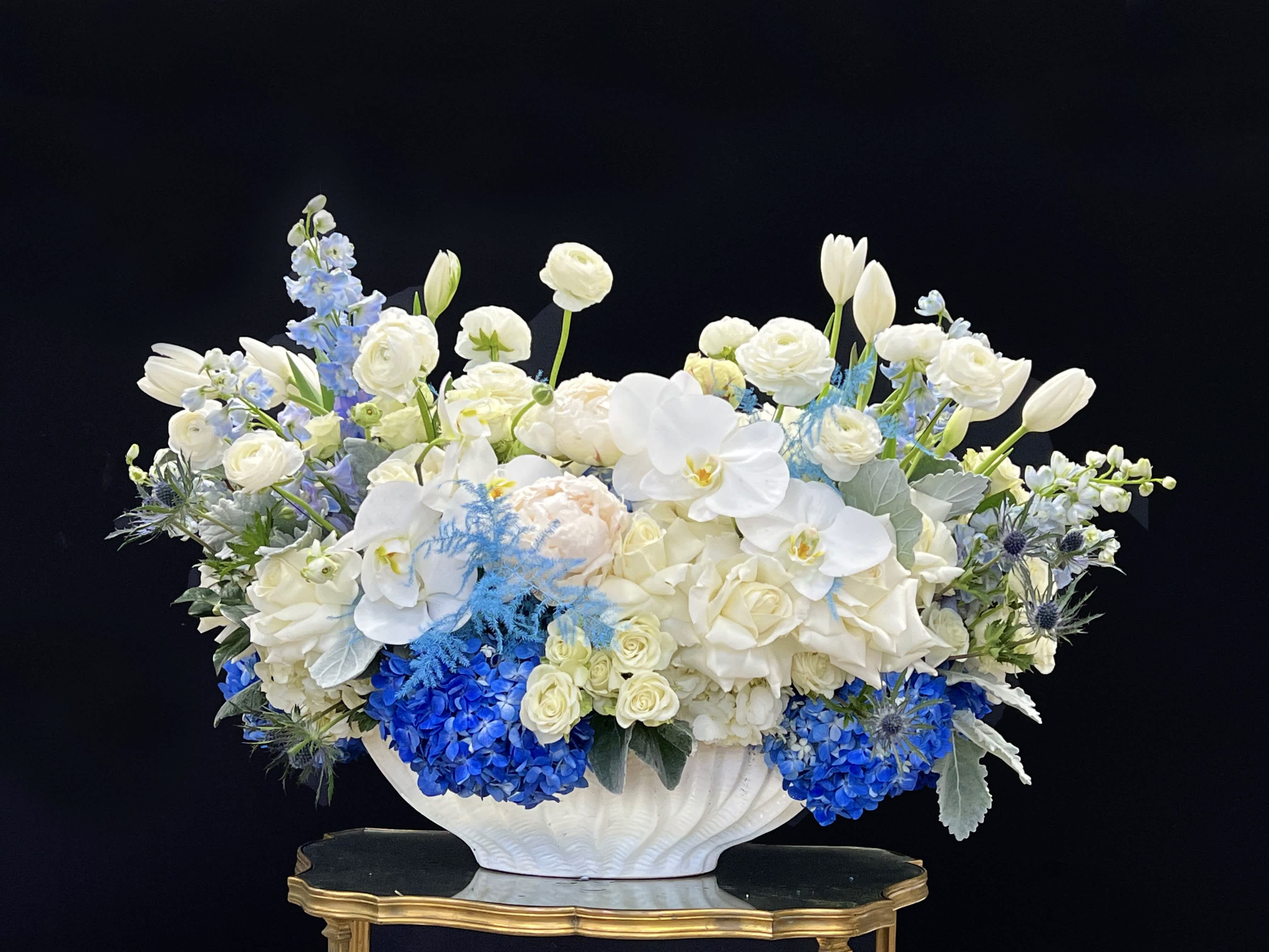 Eva’s Garden - This large ceramic vase arrangement features reflexed white Roses, Ranunculus, Tulips, blue Hydrangea, Delphinium, and Eryngium