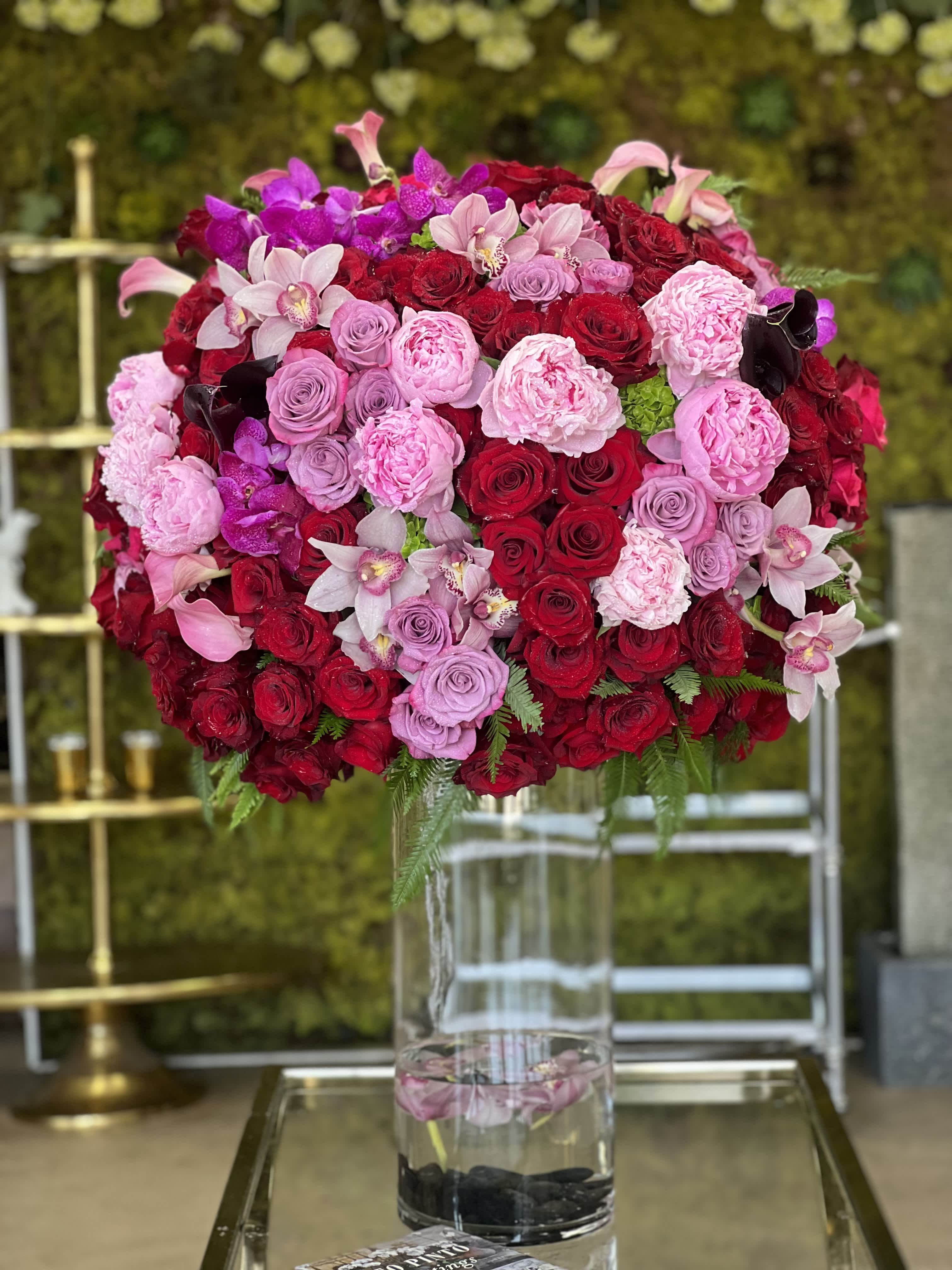 Rouge Blush - A grand arrangement with sexy and luxurious blooms... Truly a statement piece!  Featuring red and lavender Roses, pink Peonies, Calla Lilies, Cymbidium, and Vanda Orchids, set atop a tall glass cylinder vase