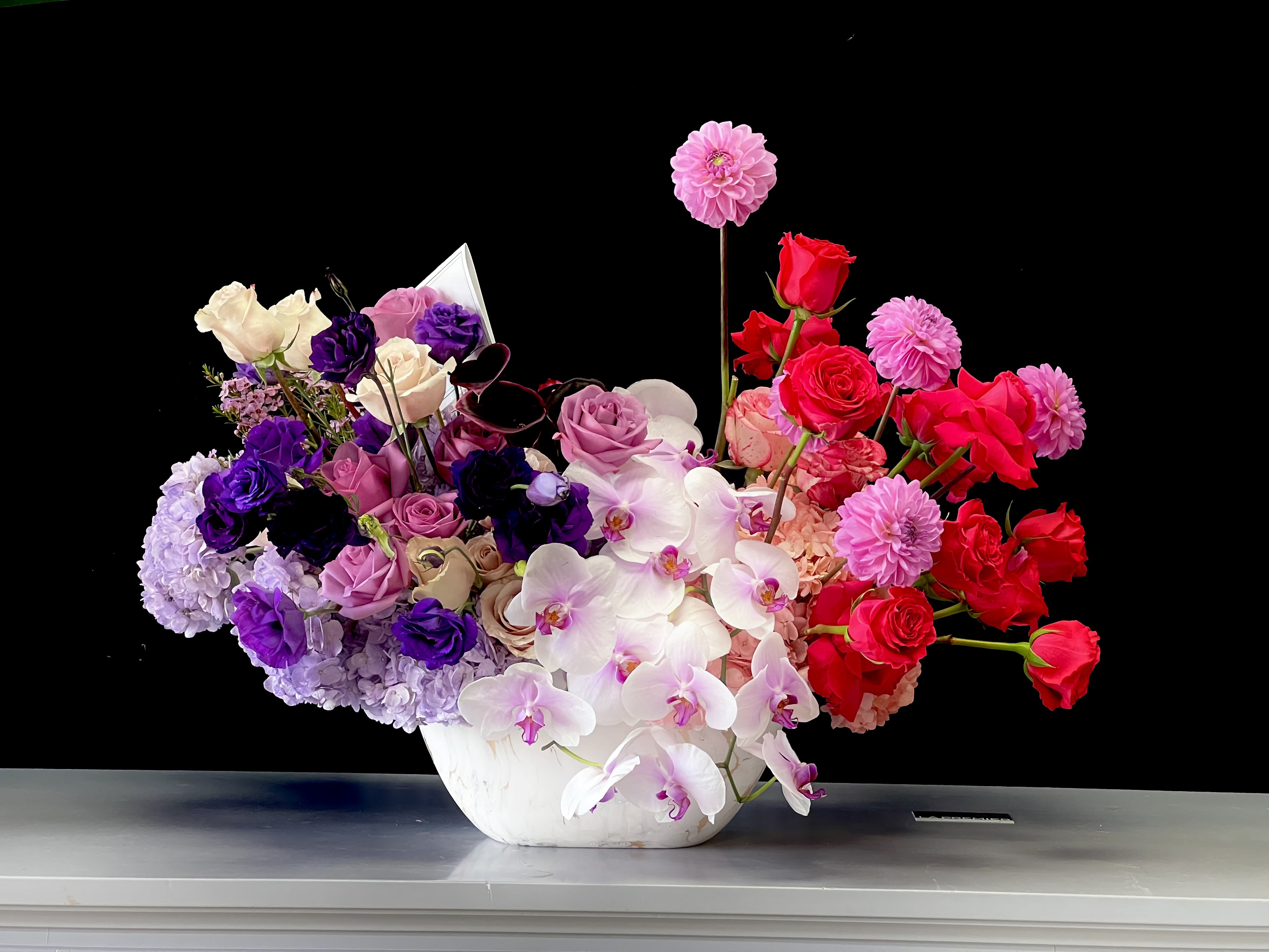 Asymmetrical bowl arrangement of pink, purple and red roses, dahlias and orchids in a white vessel.
