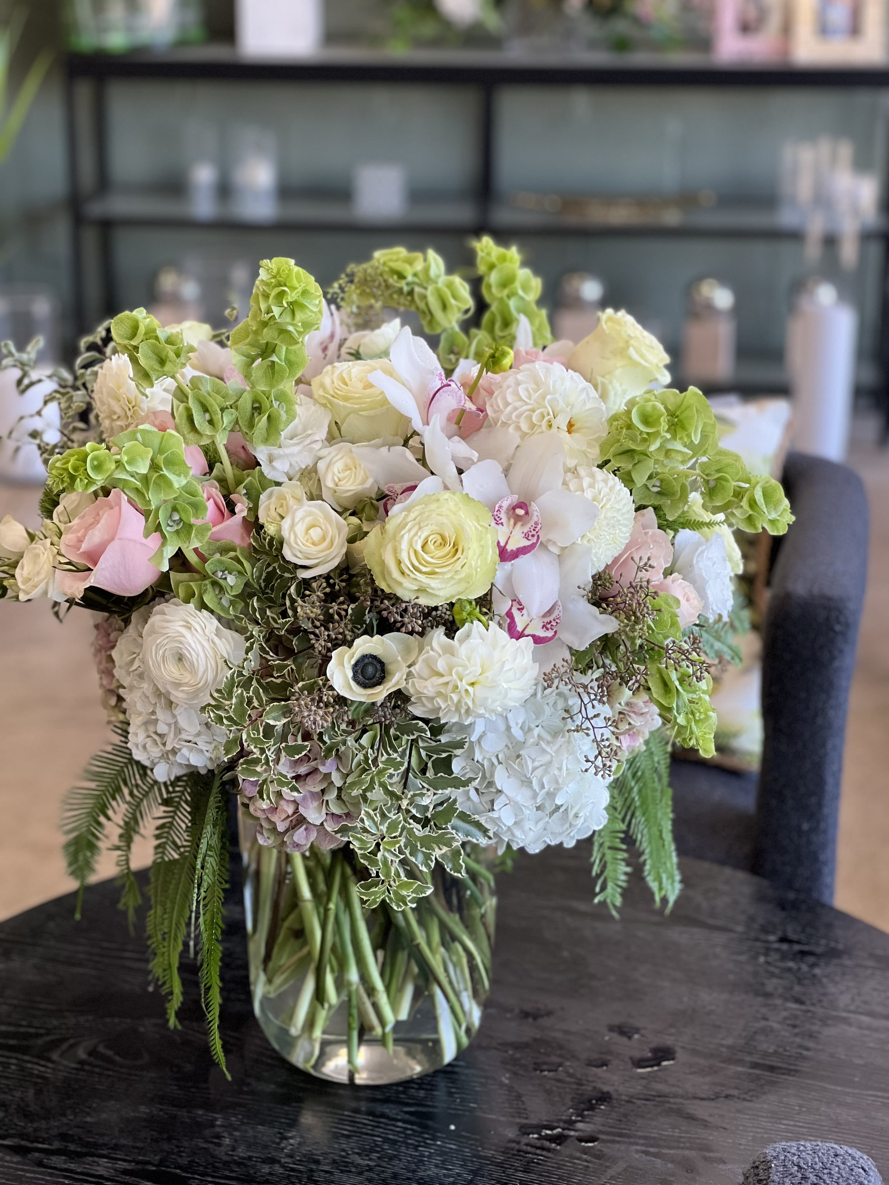 Giovana - This gorgeous cylinder vase floral arrangement features pink and white Roses, Spray Roses, Ranunculus, Bells of Ireland, Dahlias, Hydrangea, Cymbidium Orchids, and mixed greens