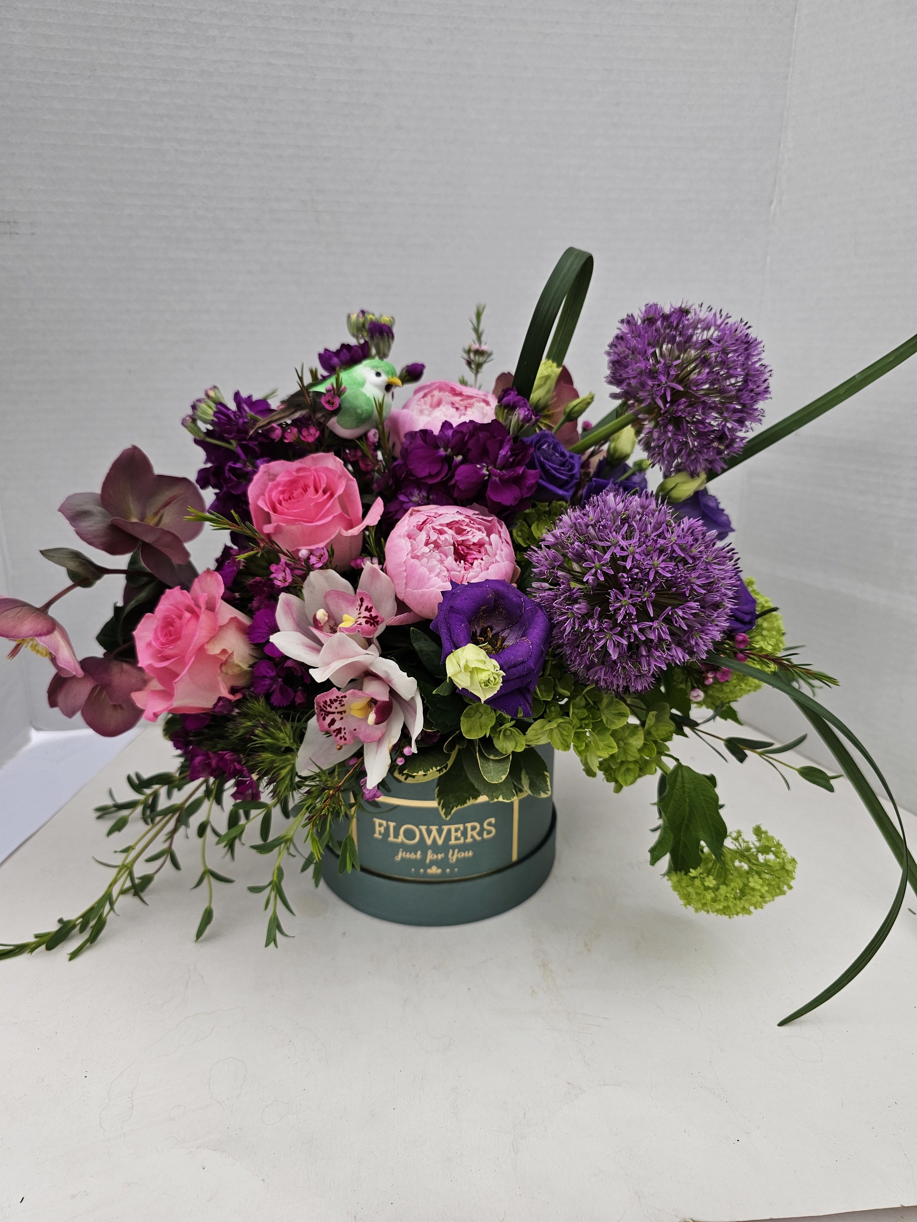 Chapeau de Fleurs - A lively hatbox arrangement that reads like a garden party in bloom-perfect as a birthday bouquet or a cheerful "just because" gift. This flower arrangement for celebrations features mixed roses and peonies alongside ranunculus and alstroemeria, delivering a bright, celebratory feel that will make the person you're thinking of feel genuinely seen and celebrated.  The design blends pink roses and rounded pink peony blooms with purple accents, spherical lavender-hued flowers, white-and-pink alstroemeria, peach roses, orange ranunculus, soft green hydrangea, and tall Bells of Ireland for vertical interest. Carnations, stock, and delicate ranunculus add texture while arched blades of grass and bright green filler bring movement and contrast. Flowers arrive arranged low and full in a round hatbox container-available in a dark green box printed with "FLOWERS" with a small decorative faux bird pick, or a soft lavender round hatbox for a gentler palette.  Ideal for birthdays, expressions of appreciation, or as a vibrant table accent, this compact arrangement works for both celebrations and everyday brightening. Sizes are presented in a generous, low-profile design that spreads outward for strong visual impact; for best longevity, keep blooms in a cool spot away from direct sunlight and change vase water if transferring to another container.