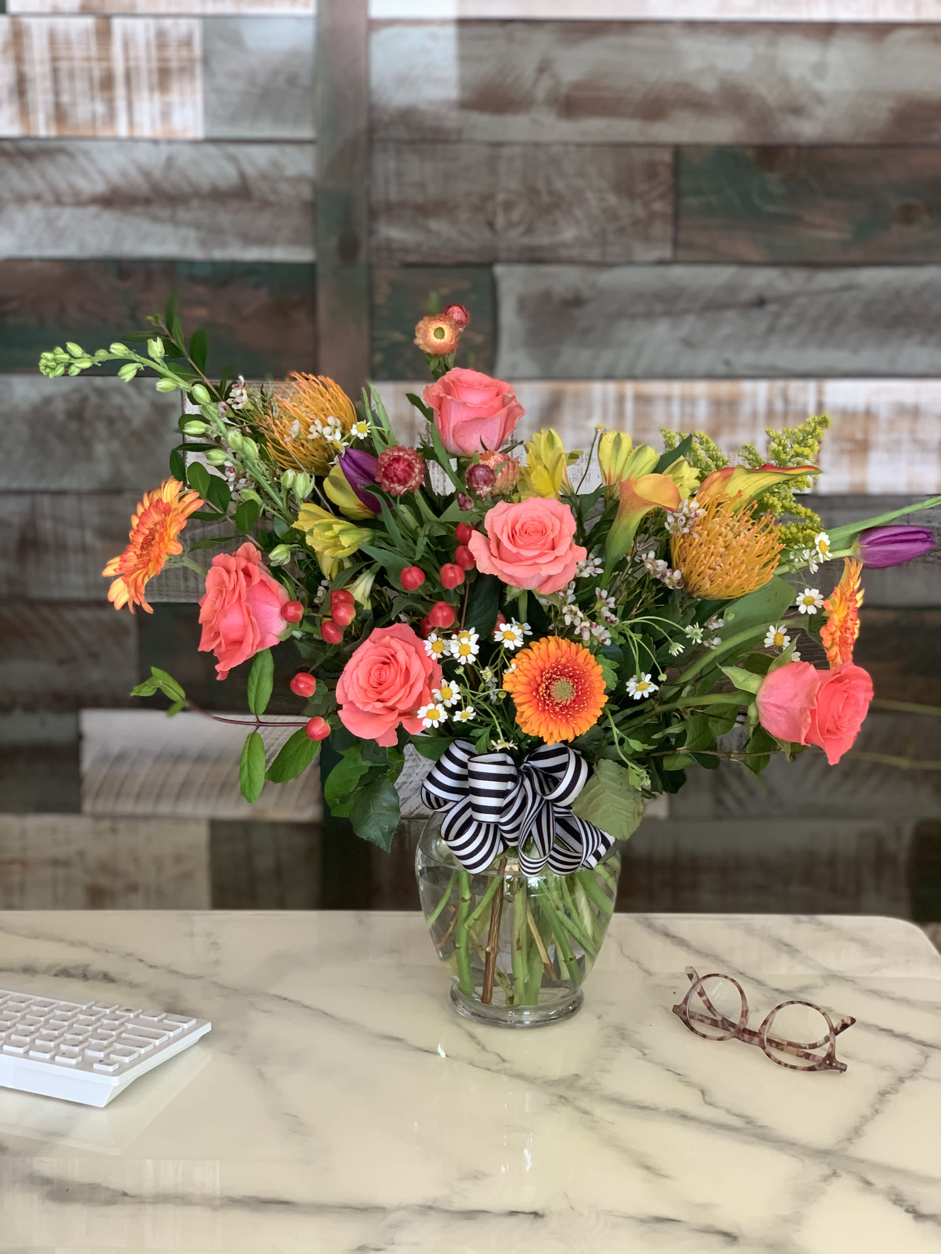Mixed bouquet of bright pink roses, orange gerberas and yellow lilies in a clear vase with striped bow on a desk