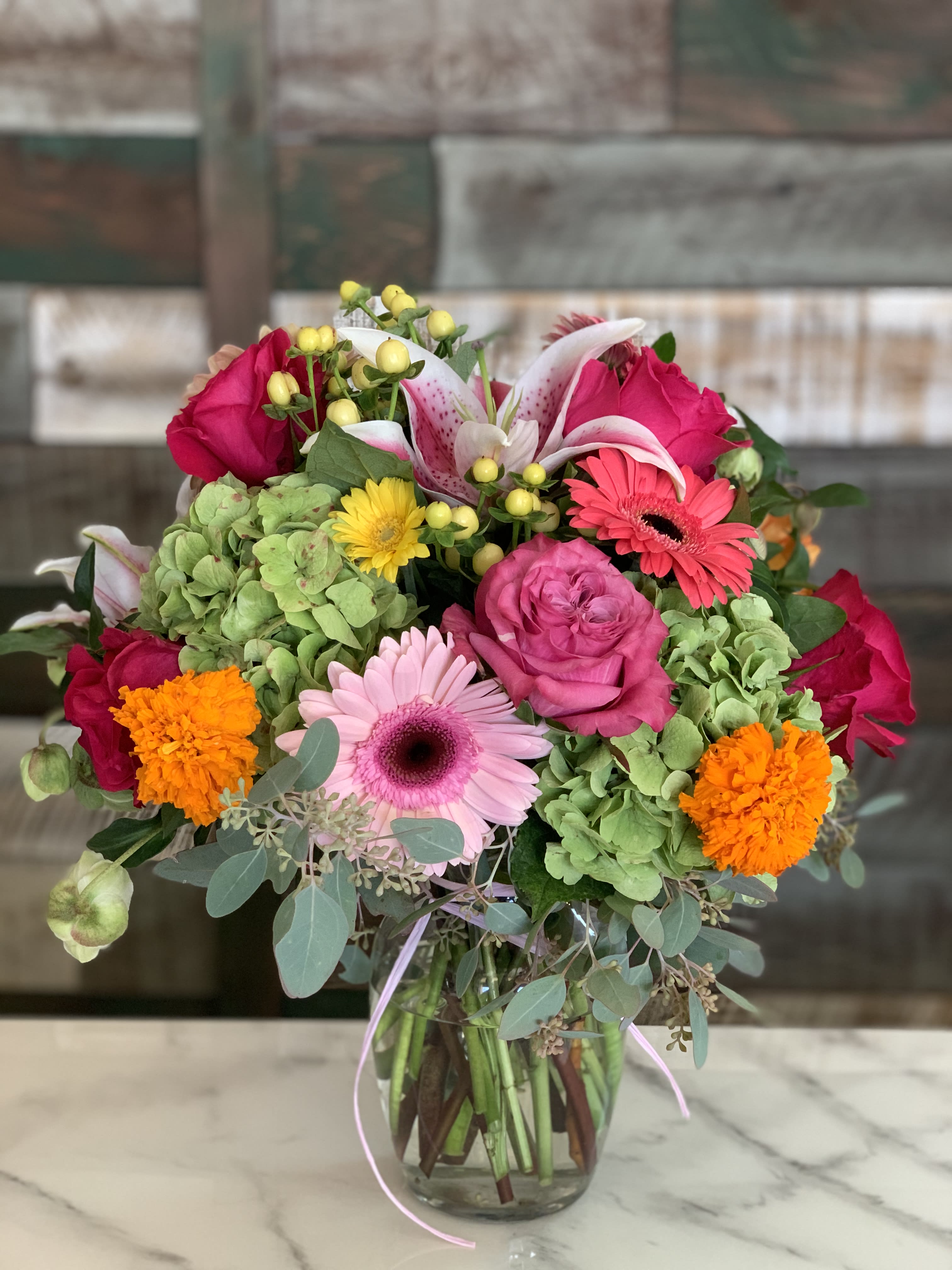Mixed bouquet of pink roses, gerbera daisies, lilies, marigolds, and green hydrangeas in a clear glass vase