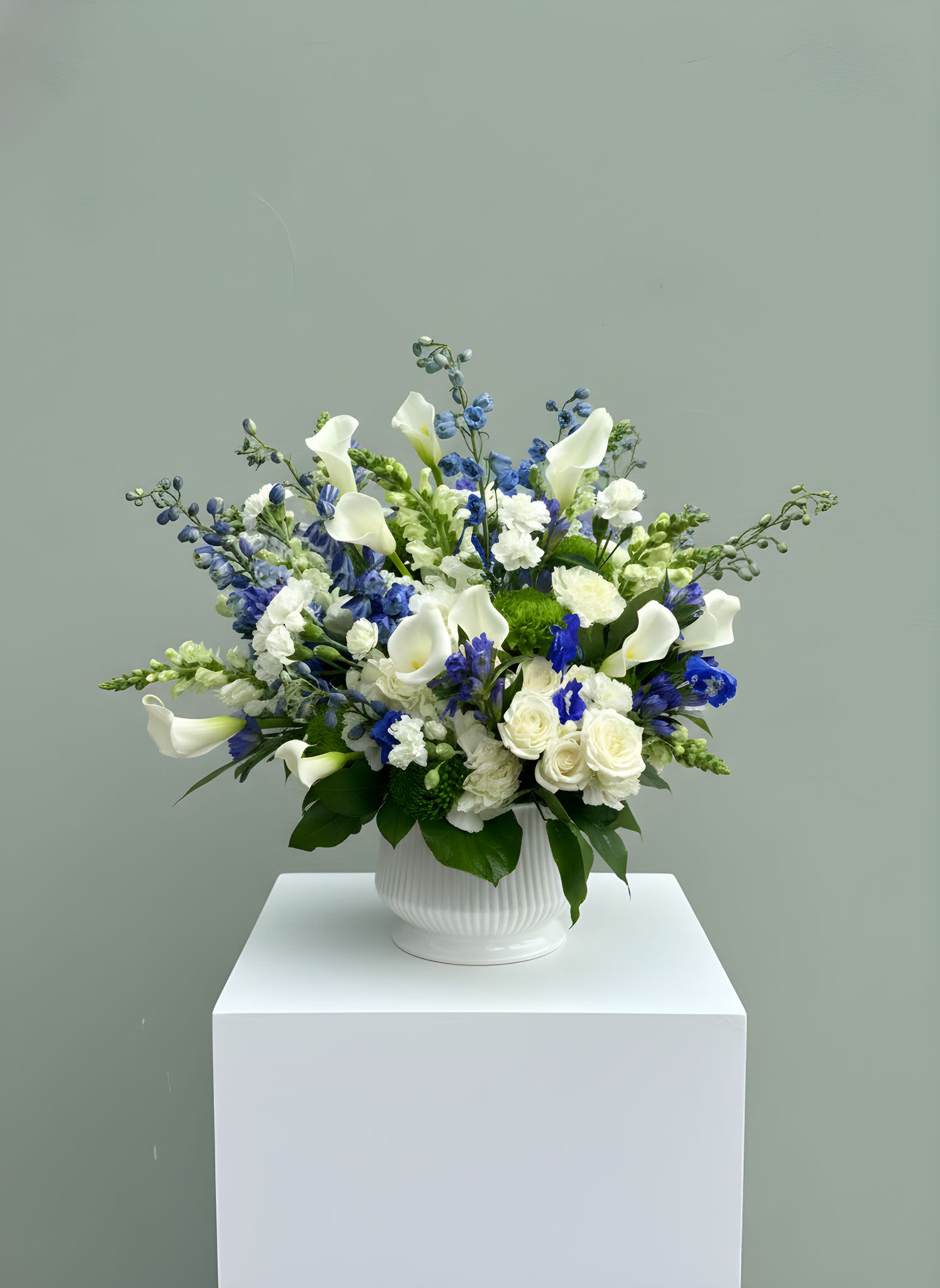 Azure Elegance Floral Arrangement - A refined and timeless floral arrangement featuring a stunning blend of crisp white blooms and rich blue accents. Designed with elegant calla lilies, premium white roses, delicate delphinium, and lush seasonal textures, this arrangement creates a fresh, serene, and sophisticated look.  Set in a modern ceramic vase, this design is perfect for expressing sympathy, celebrating milestones, or adding a touch of calm luxury to any space  Please note: Flower varieties &amp; greenery fillers may vary slightly based on availability while maintaining the same color palette, style, and overall fullness of the arrangement.