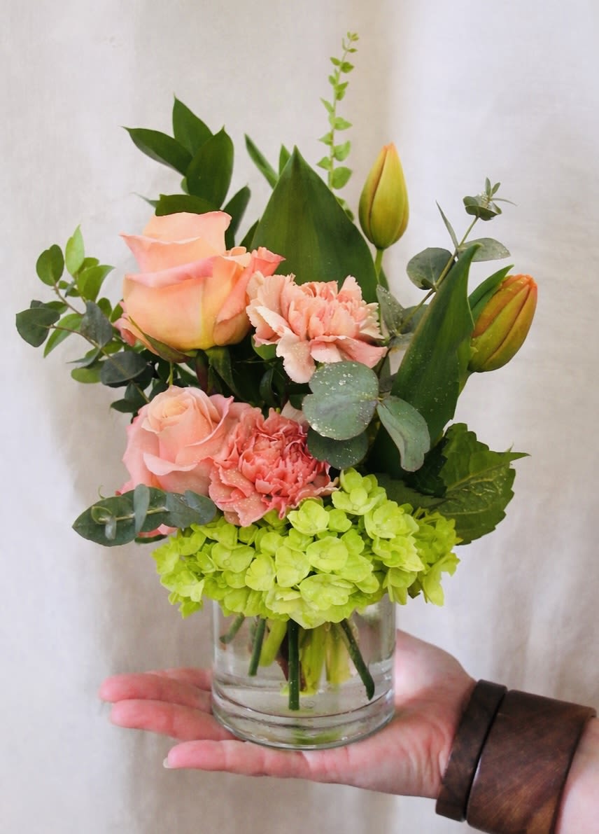 Mini Golden Hour  - A bright, cheerful hand-held arrangement with peach roses, soft peach carnations, orange tulips, and fresh green hydrangea. Styled in a small glass vase with clean greenery for a fresh, modern look.